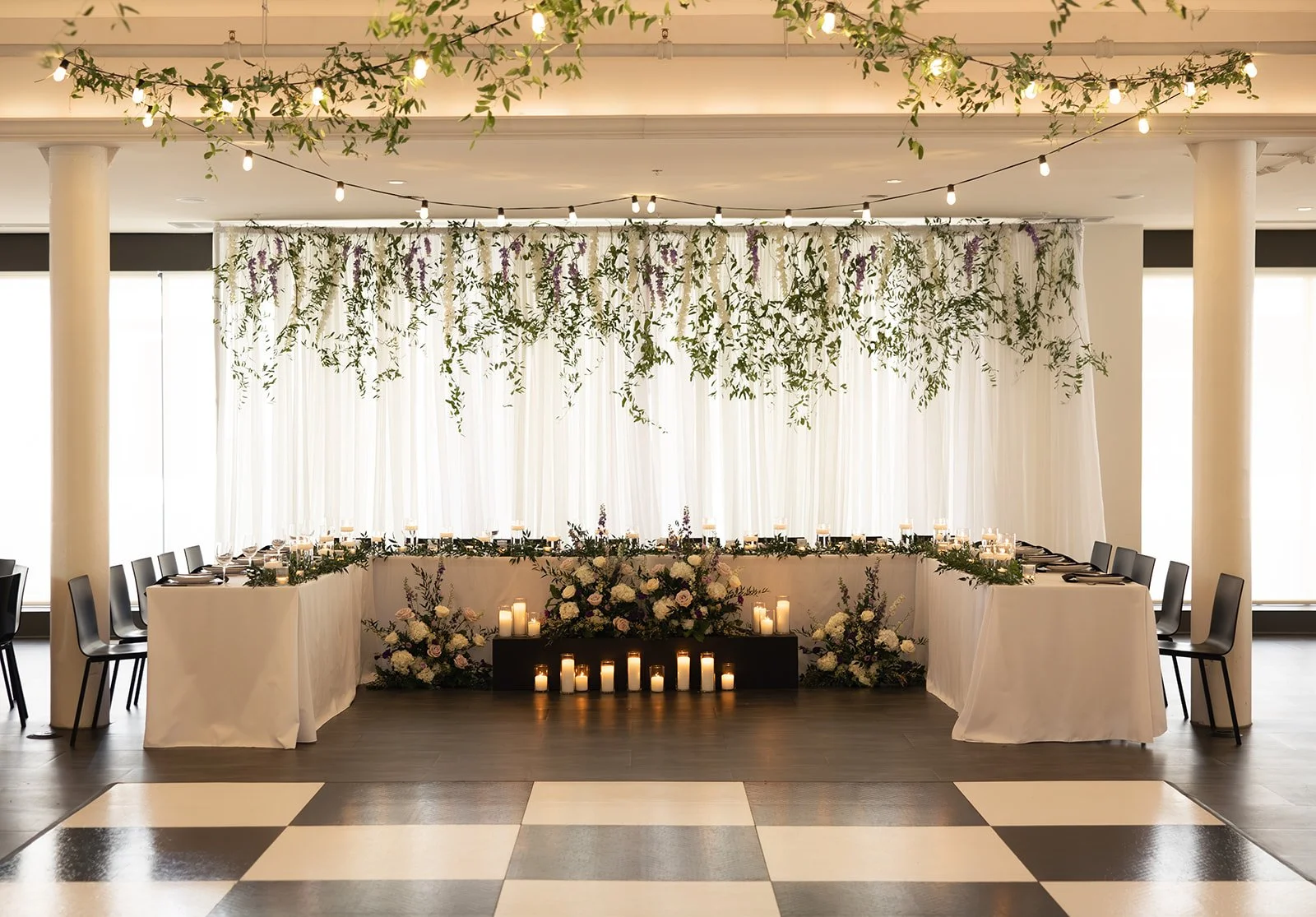 Elegant wedding reception setup with a long U-shaped table decorated with white tablecloth, candles, and floral arrangements, featuring hanging greenery and purple flowers above, and string lights creating a warm ambiance.