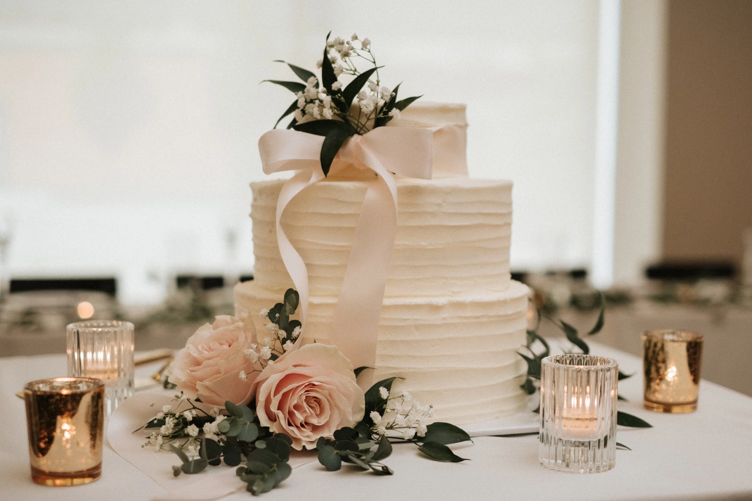 A wedding cake decorated with flowers and a pink ribbon, surrounded by pink roses and candle holders.