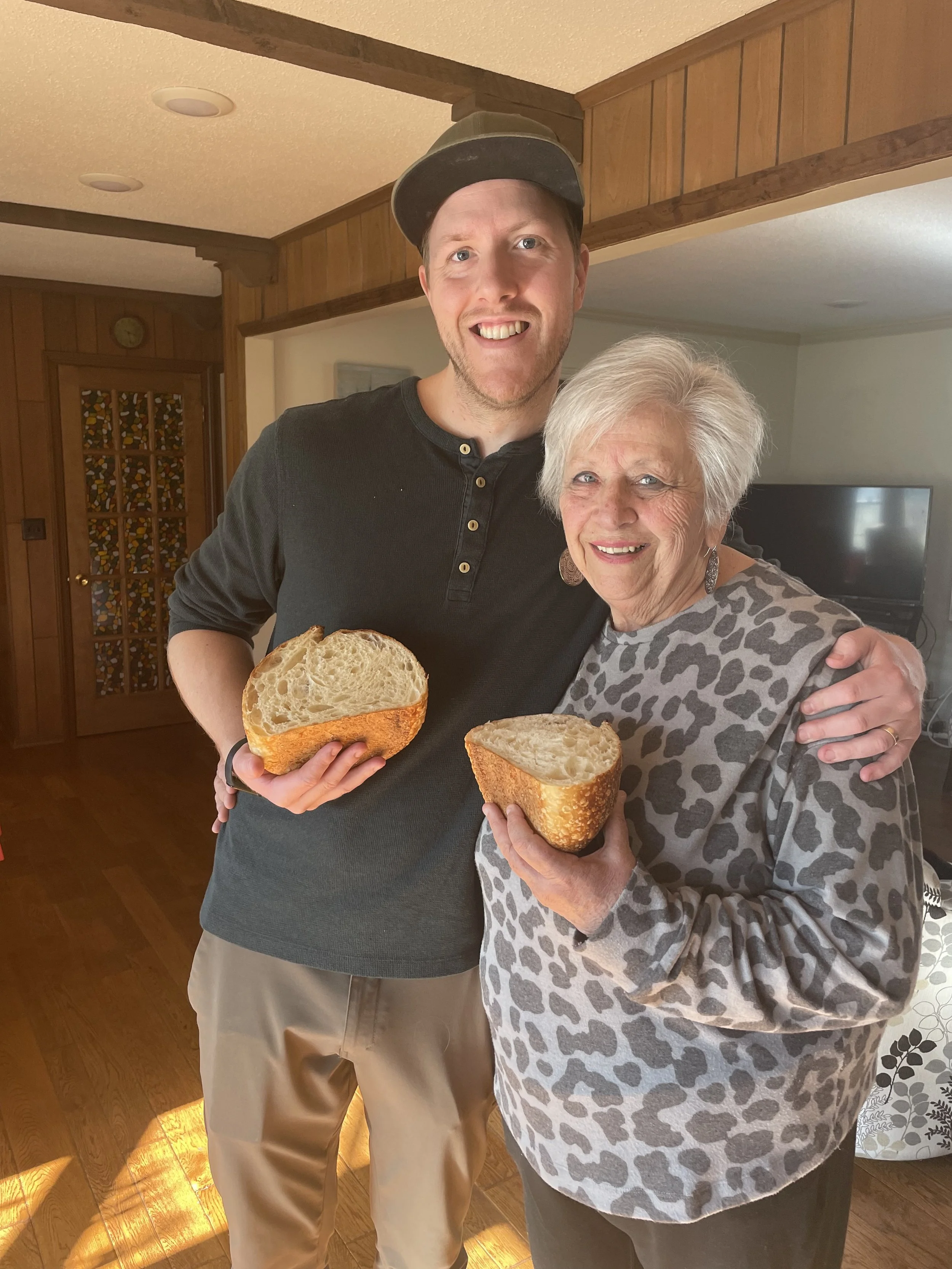 A young man and an elderly woman are smiling and holding large slices of bread indoors. The young man is wearing a black long sleeve shirt and a hat, while the woman is wearing a leopard print sweater.