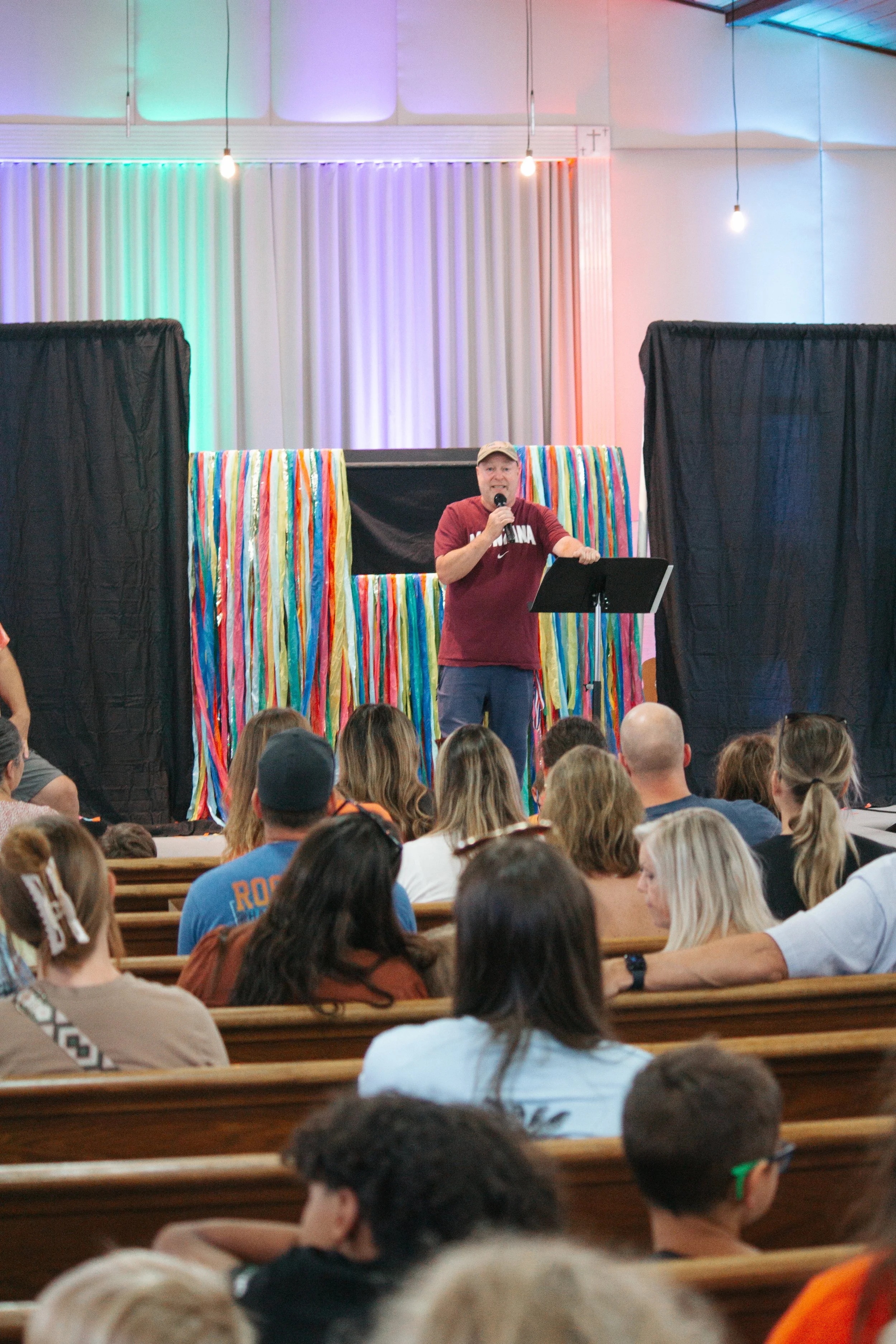 A man wearing a tan cap and a maroon shirt stands on a stage with colorful streamers, speaking into a microphone to an audience seated on wooden pews.
