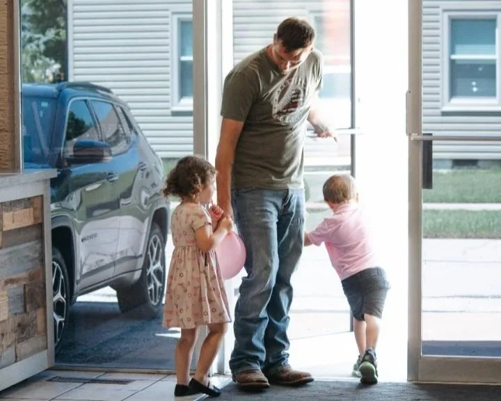 Father with two children at a doorway, one girl holding a pink balloon and an older boy near the door.