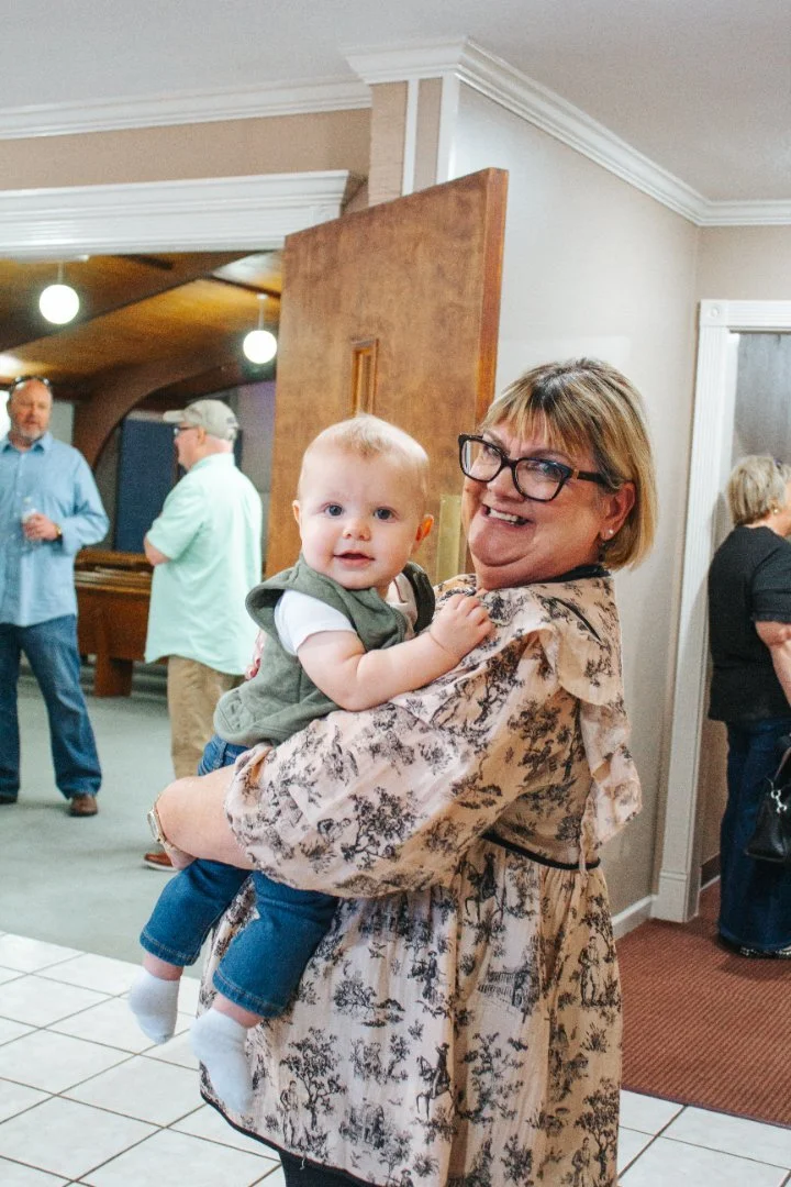 A woman holding a baby in a room with several other people in the background, some standing and talking, a pool table visible, and a partially open wooden door.
