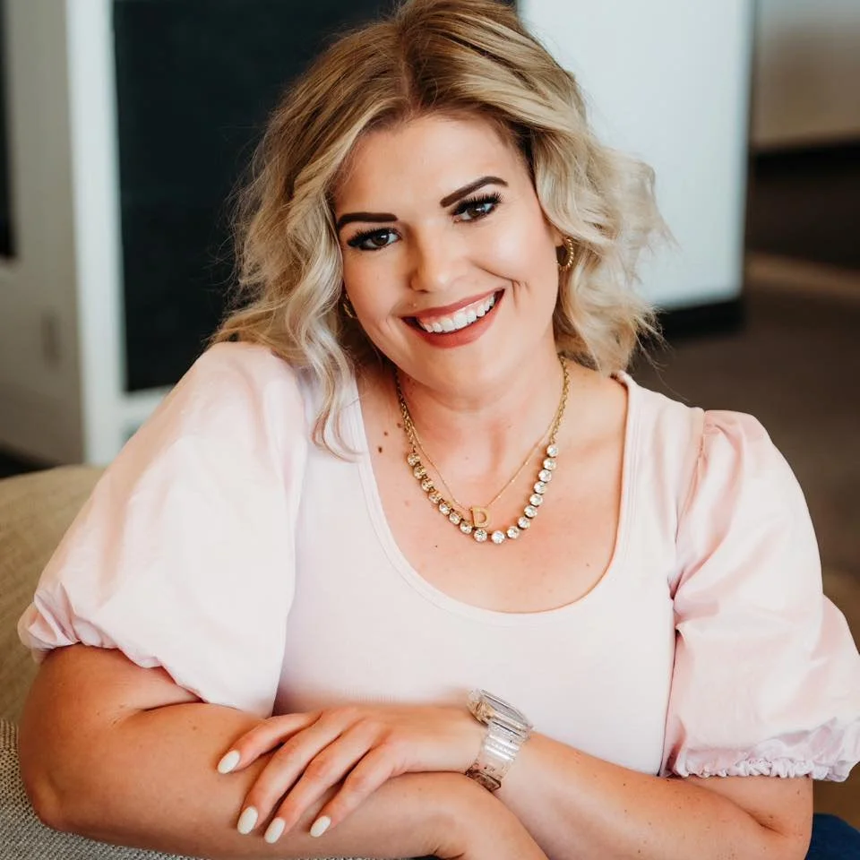 A smiling woman with blonde, curly hair wearing a pink puff-sleeve top, gold jewelry including a necklace with a 'D' pendant, earrings, and a watch, sitting indoors with a blurred background.