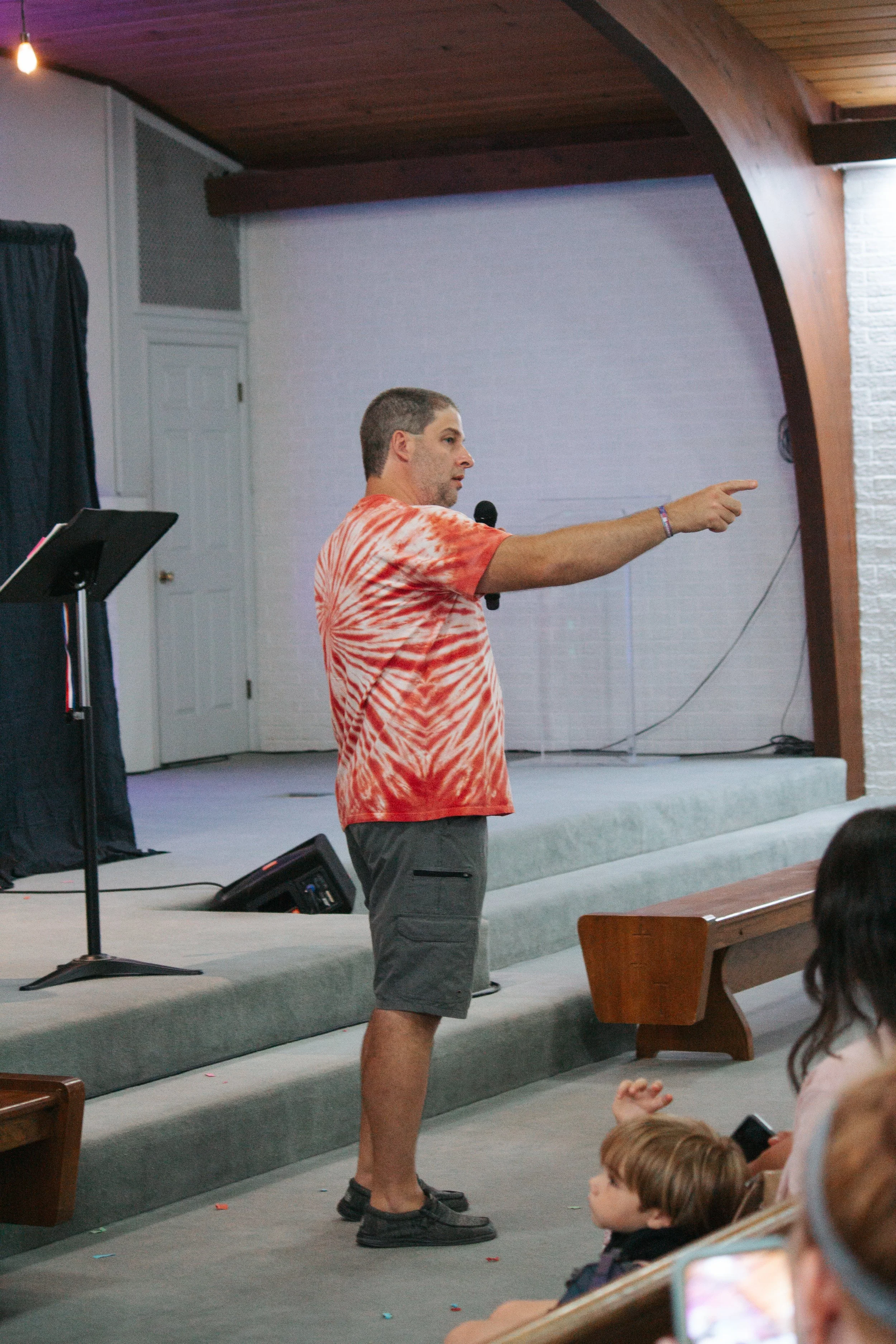 A man in a tie-dye shirt and gray shorts stands on a stage, speaking into a microphone and pointing to the right. The stage has a music stand and a black speaker; the background includes a white wall with a door and a wooden arched structure.