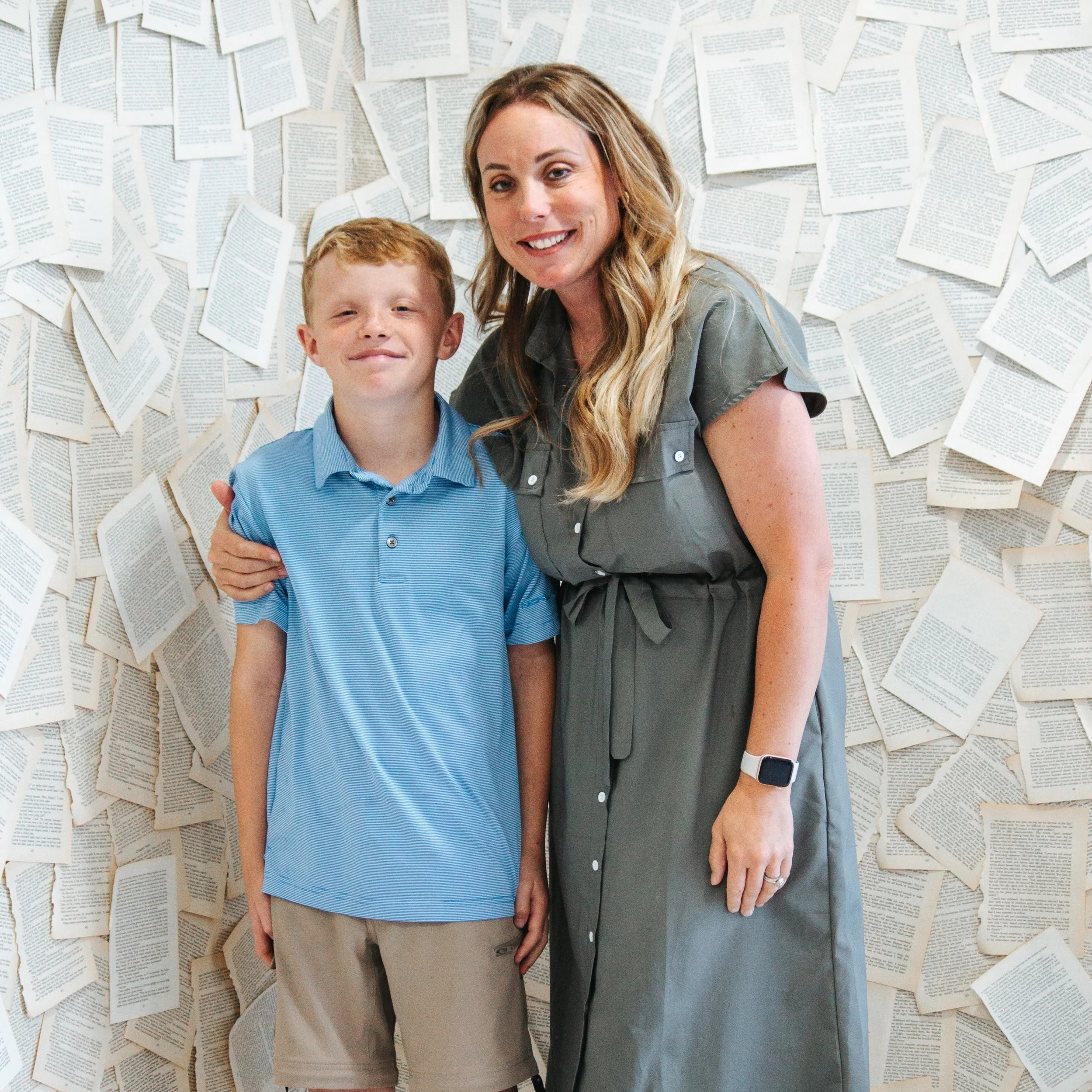 A woman and boy standing together in front of a wall covered with open books, smiling at the camera.