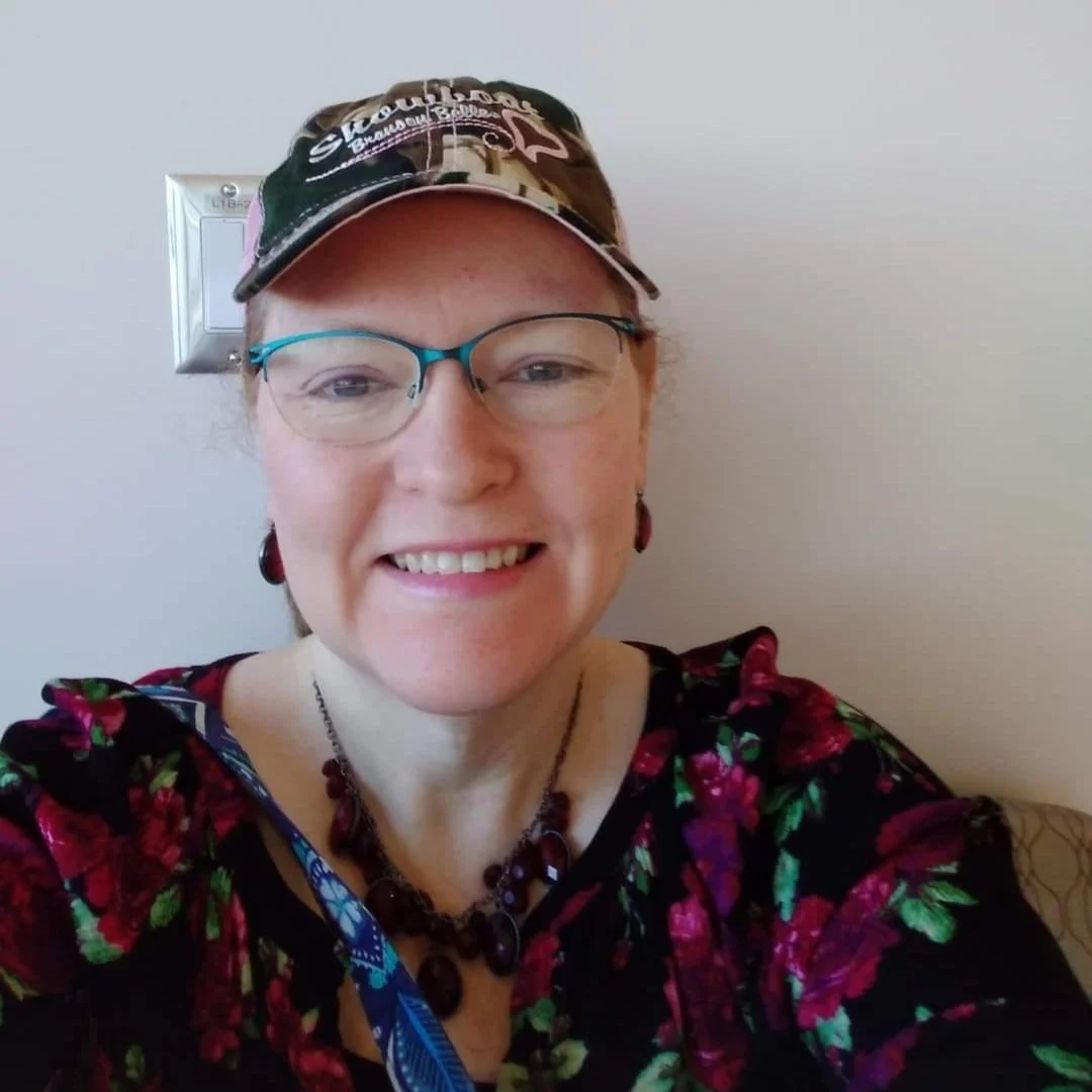 A woman with glasses, wearing a camo cap, earrings, and a floral top, smiling indoors.