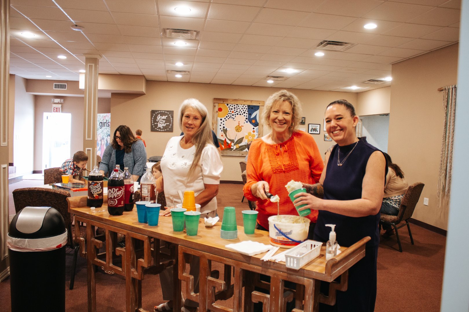 Three women standing behind a table with cups and drinks, serving ice cream on a casual indoor gathering. Other people are seated at tables in the background.