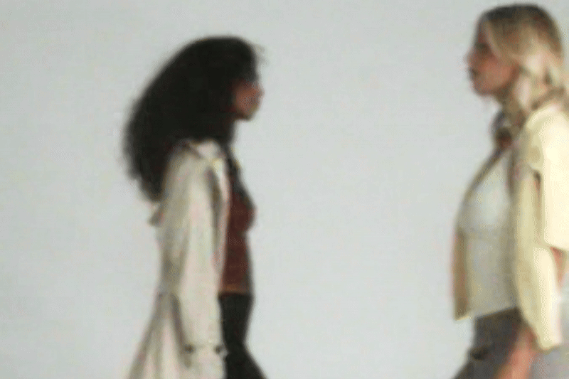 Two women standing face-to-face indoors against a plain white wall.