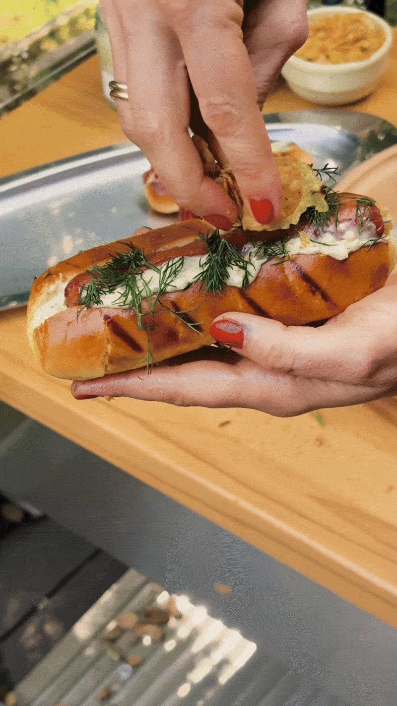 A person assembling a hot dog sandwich with condiments and herbs on a wooden table.