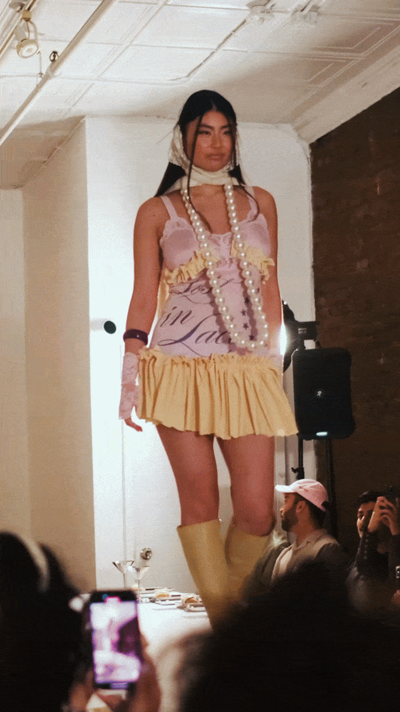 A woman walking on a runway in a pastel-colored outfit with large pearl necklaces, a bandana, and yellow knee-high boots, during a fashion show.