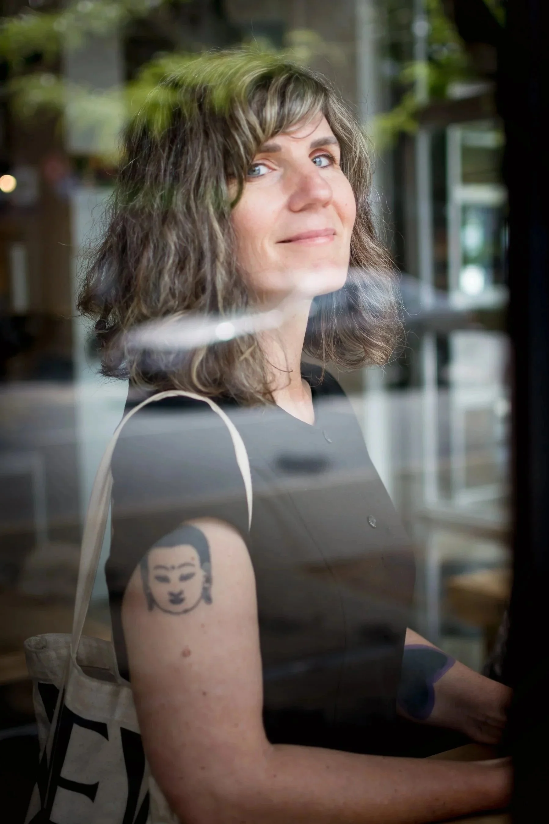 Roberta McDonald looking out from a coffee shop window with an engaging smile. She has multiple tattoos and is carrying a tote bag