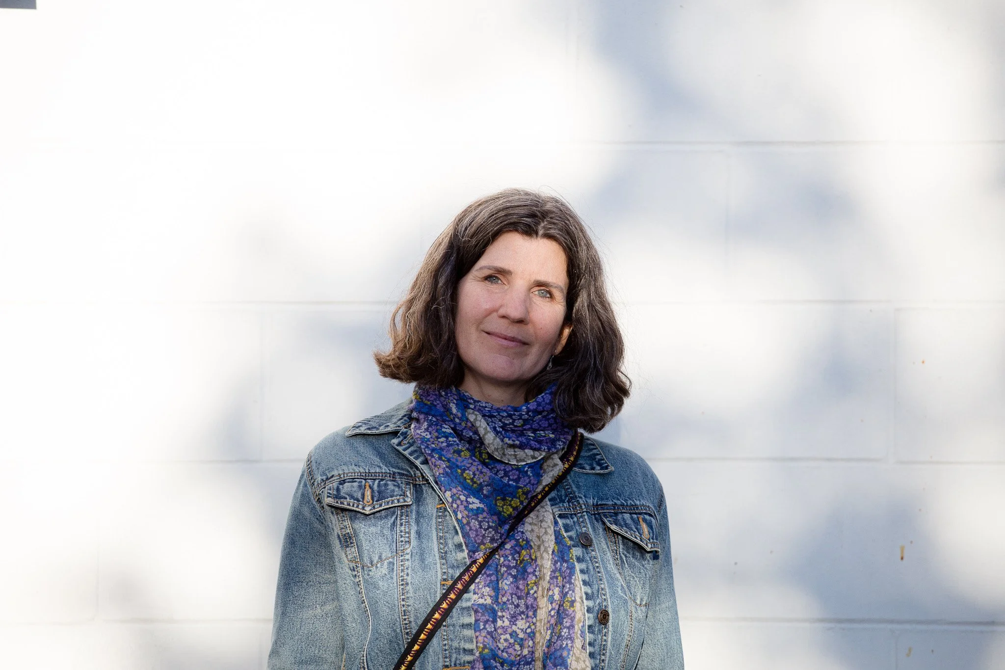 Roberta is standing in front of a white wall with bokeh shadows, wearing a jean jacket and scarf. Part of an ADHD portrait project by Ryan Walter Wagner