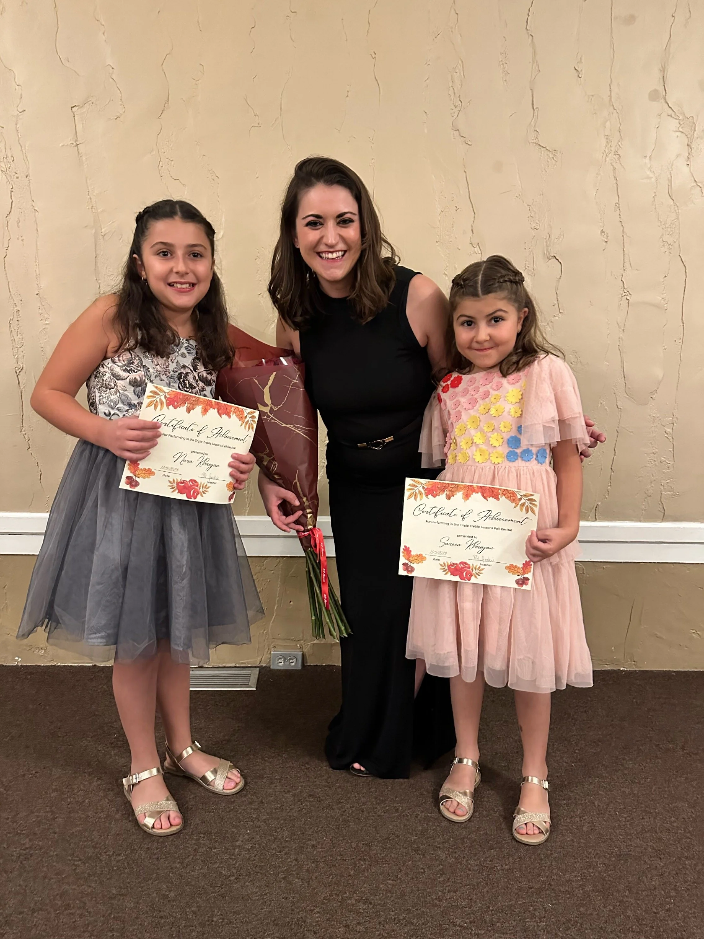 Two young students holding certificates and Jackie in a black dress standing between them, in an indoor setting with beige textured walls.