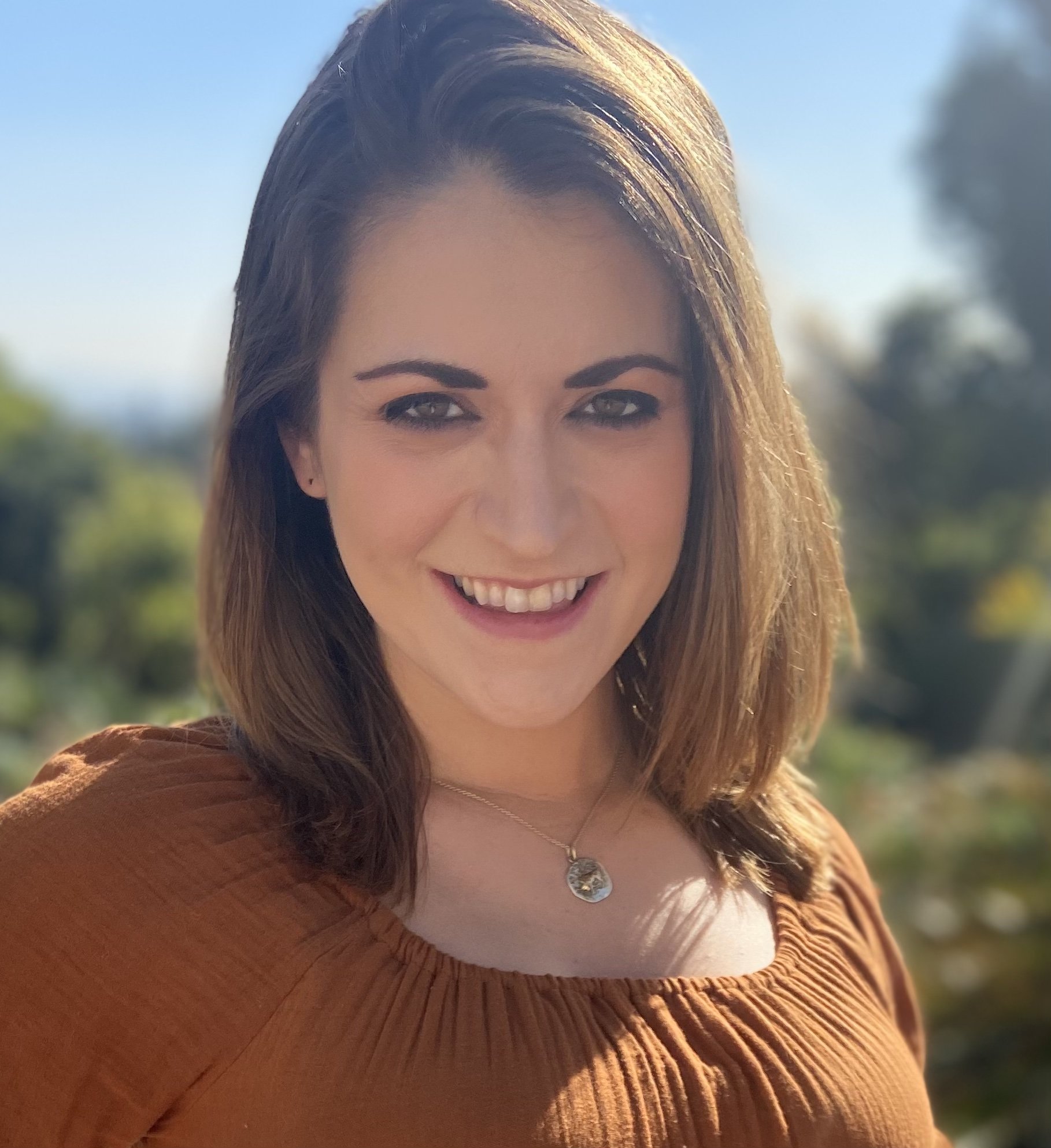 A smiling woman with shoulder-length brown hair standing outdoors with a blurred natural background.