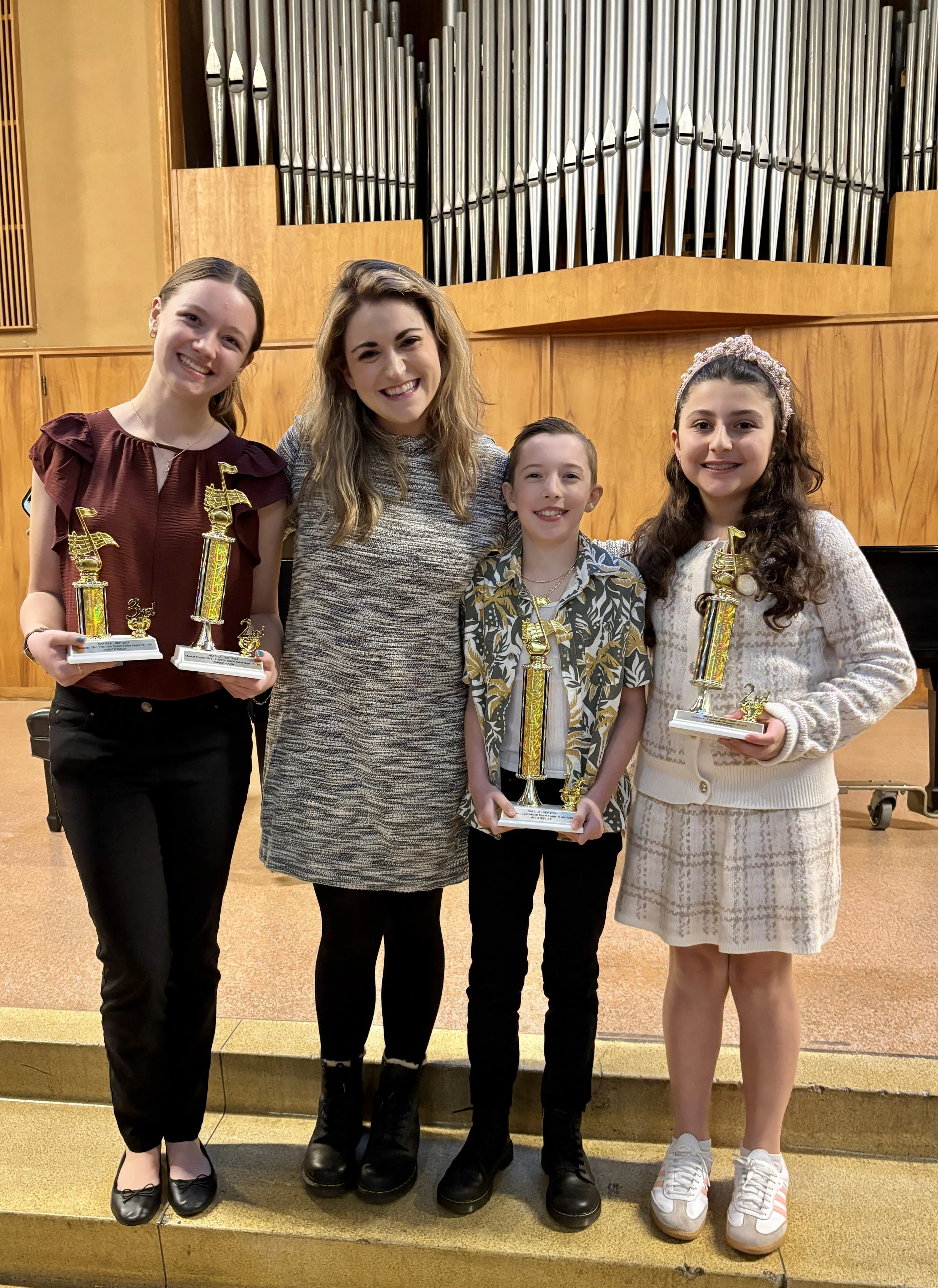 Four young students and their teacher standing on a stage, holding trophies, in front of a large pipe organ in a concert hall.
