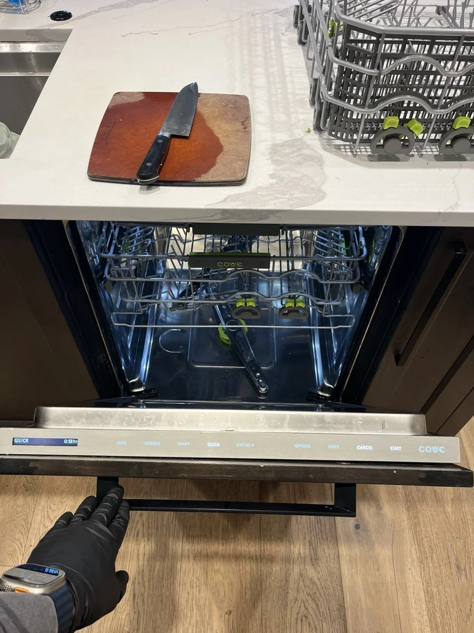 Open dishwasher with a black-handled kitchen knife on a wooden cutting board on the adjacent counter. The dishwasher is empty and turned on, with the control panel showing a cycle in progress.