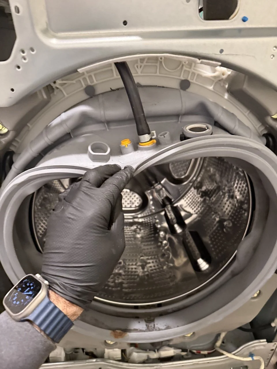 A person wearing black gloves and a smartwatch is opening the door of a washing machine, revealing the drum inside.