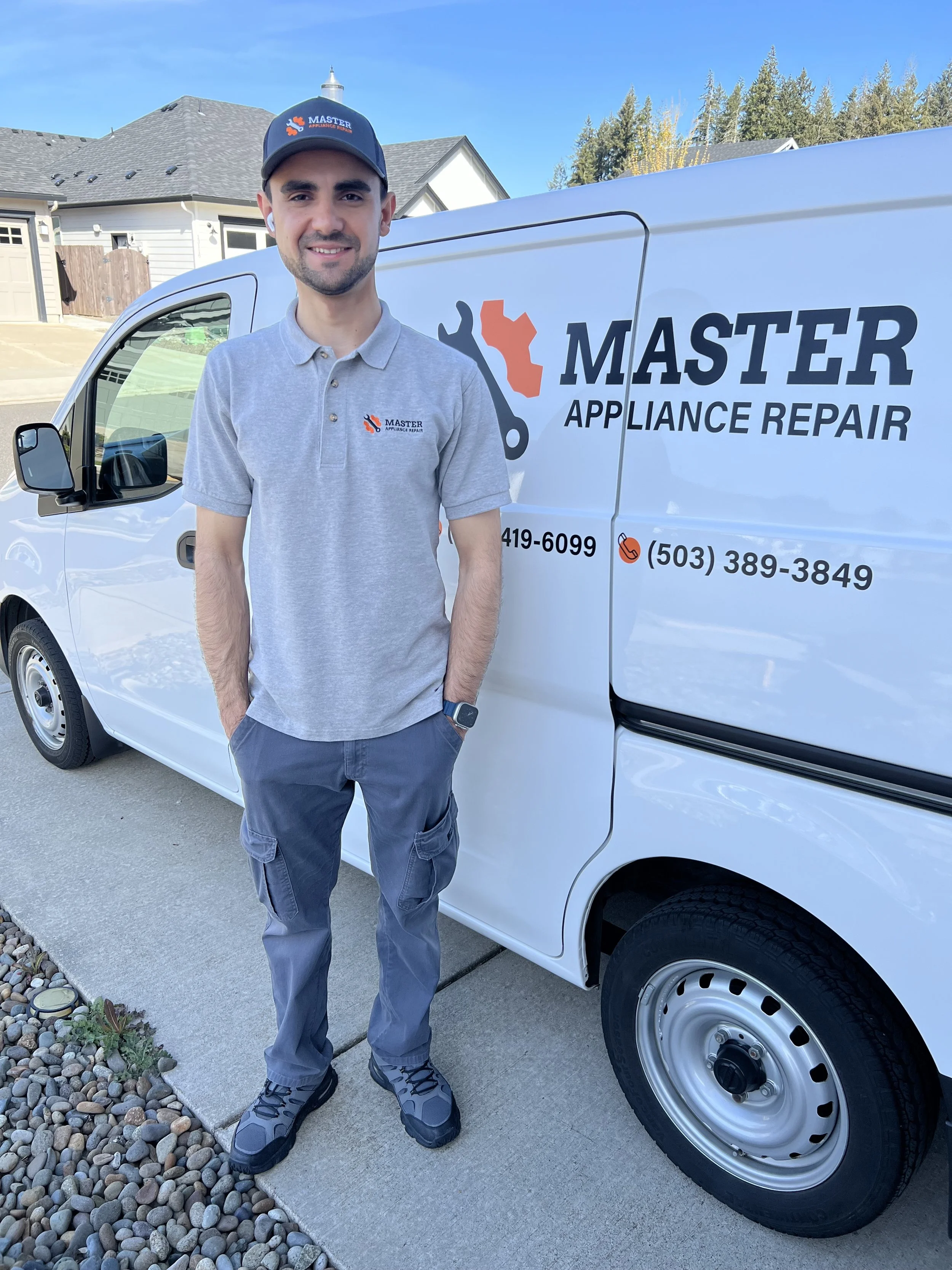 A young man wearing a gray polo shirt, gray pants, and sneakers standing next to a white van with the logo and contact information for Master Appliance Repair. He is smiling and wearing a black cap with the company's logo, and has earbuds in his ears.