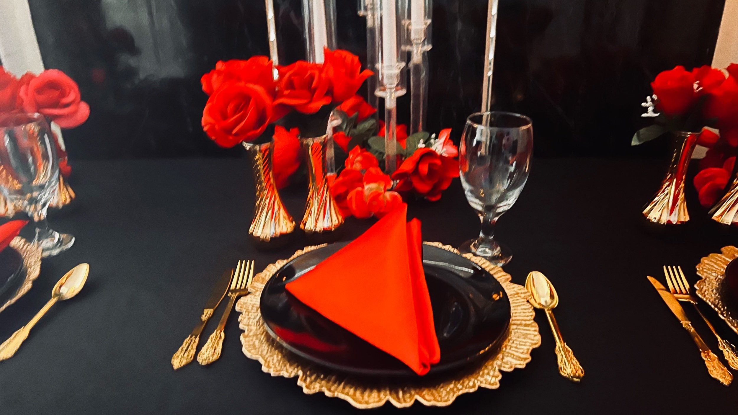 A table set for a celebration with a black and gold theme, featuring red flowers, gold utensils, a gold placemat, a black plate with an orange napkin, and gold party hats with red accents, against a black tablecloth background.