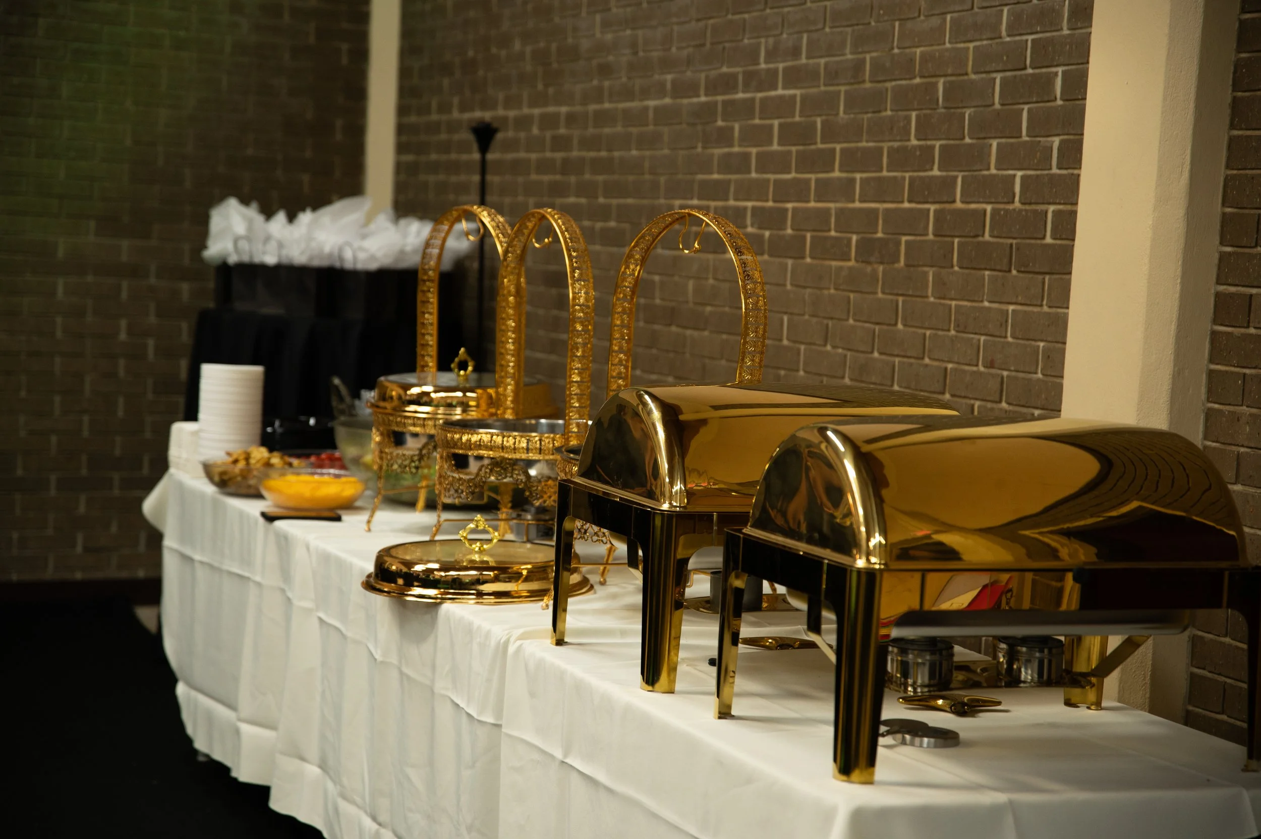 Buffet table with gold chafing dishes, serving trays, and food dishes, set against a brick wall, with stacked white plates and bowls on the side.