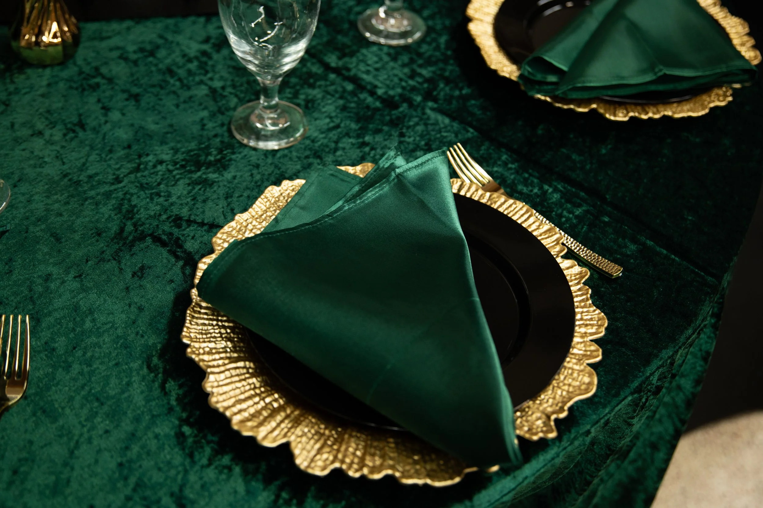 A formal table setting with a green napkin folded on a black plate, gold-colored charger plate, gold fork on the right side, and clear glassware on a dark green tablecloth.