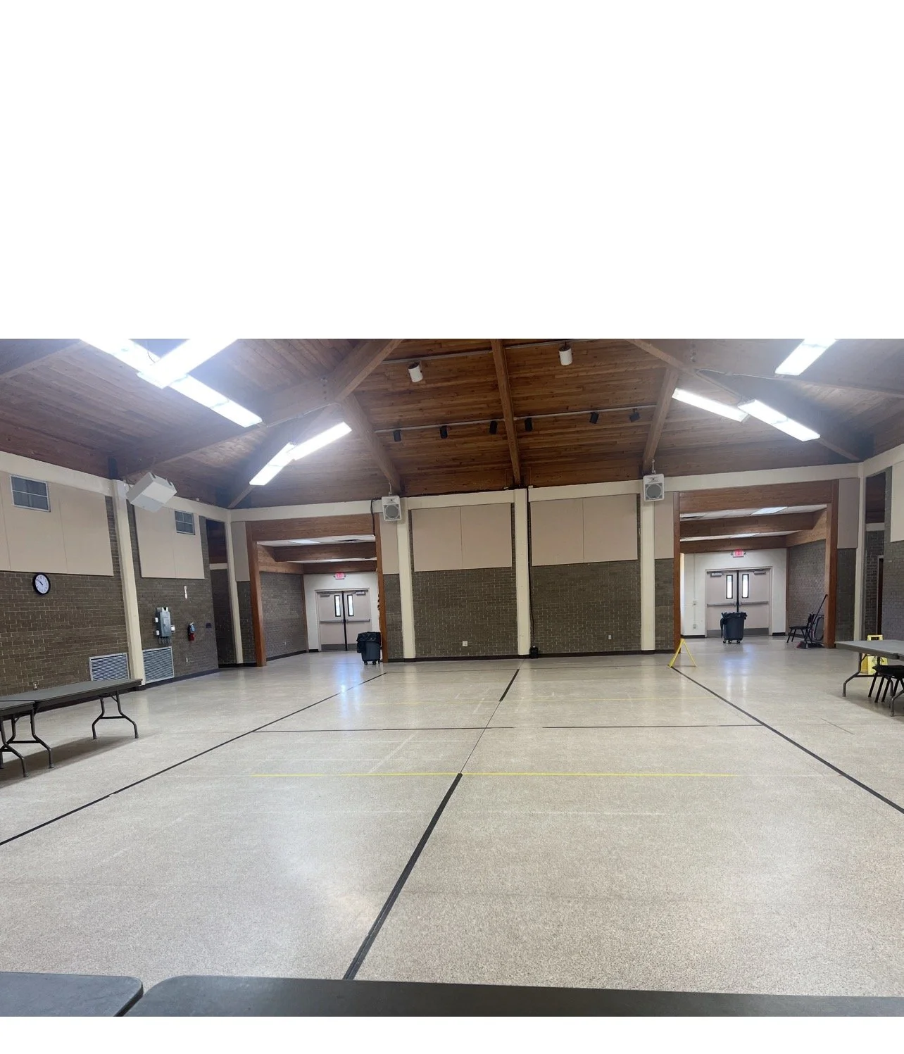 Empty multipurpose room with high wooden ceiling, fluorescent lighting, brick and beige walls, folding tables against the sides, and a basketball court outline on the floor.