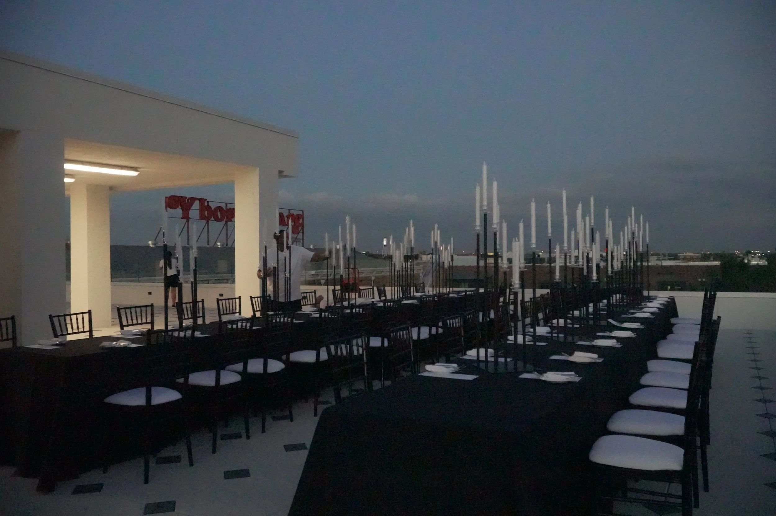 Rooftop restaurant dining area set up with black tables and white chairs, with a city skyline in the background during dusk.