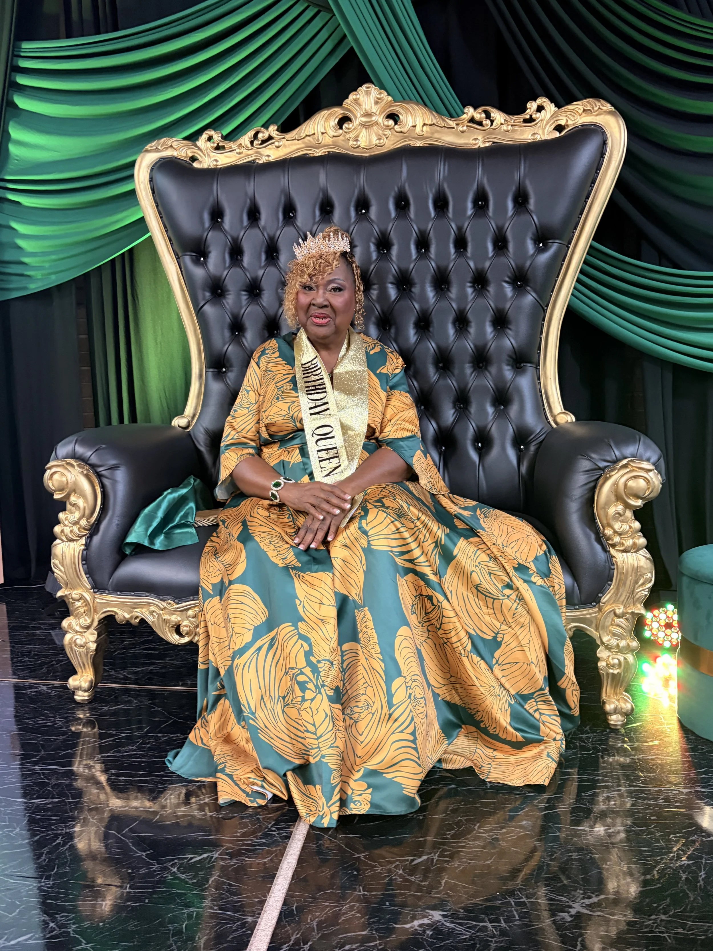 A woman sitting on a large black and gold throne with a green and black draped curtain backdrop. She is wearing a crown, a sash reading 'Birthday Queen,' and a flowing dress with green and gold leaf patterns.