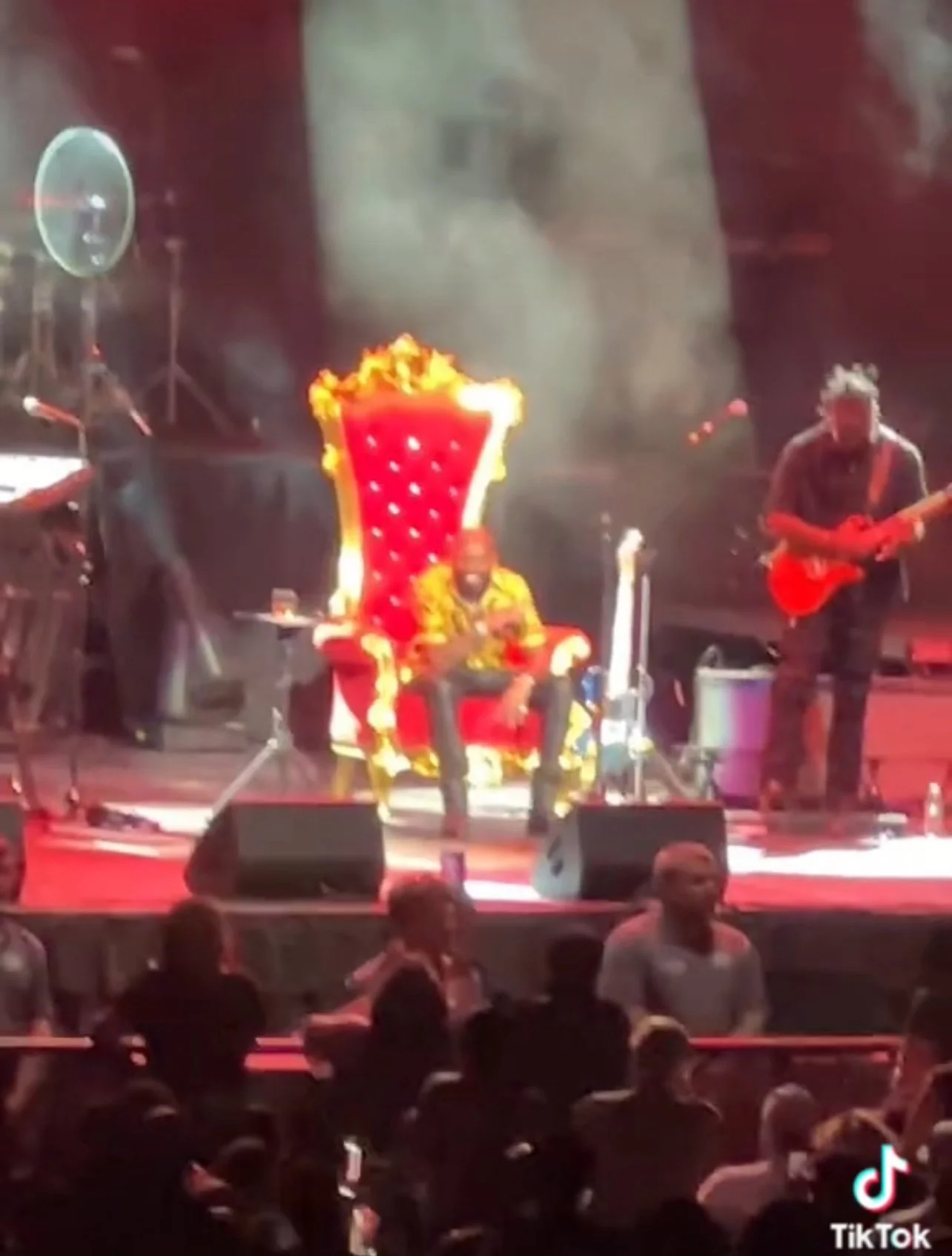 Performer sitting on an ornate red and gold throne on stage, playing a musical instrument, with a guitarist on the right and an audience in front.