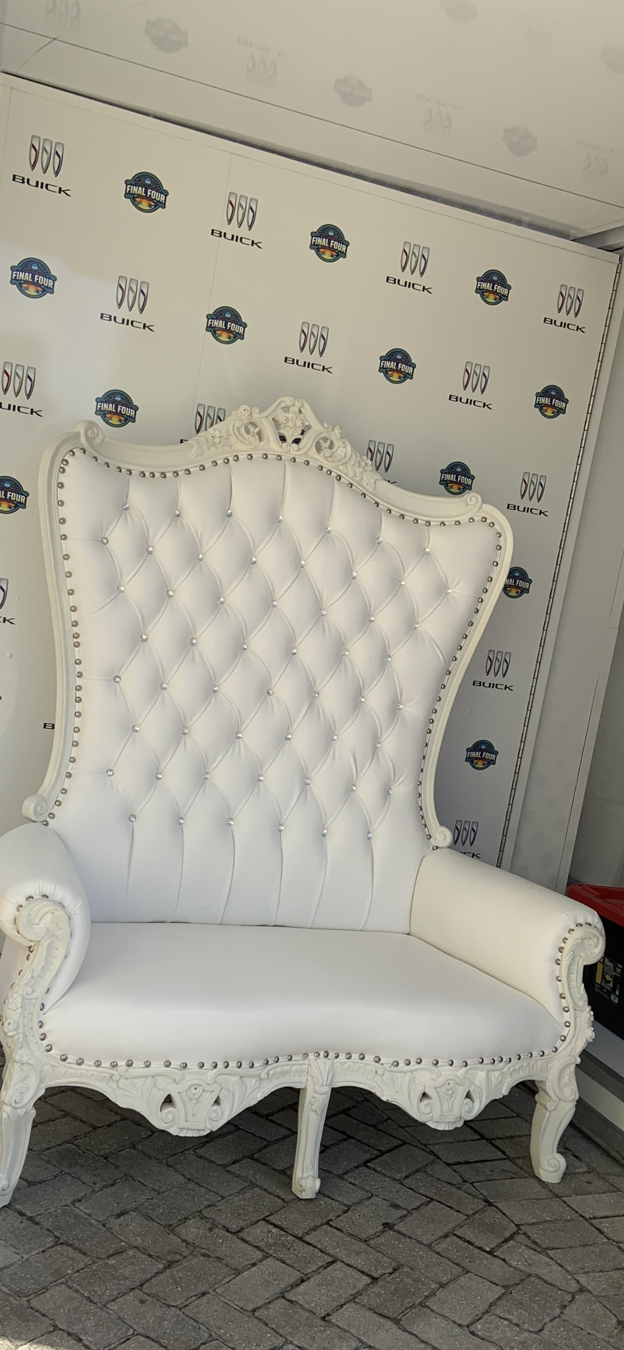 A white, antique-style high-back armchair with ornate carvings, button-tufted upholstery, and nailhead trim, placed on a brick pavement with a backdrop featuring logos of Buick, Final Four, and other sponsors.