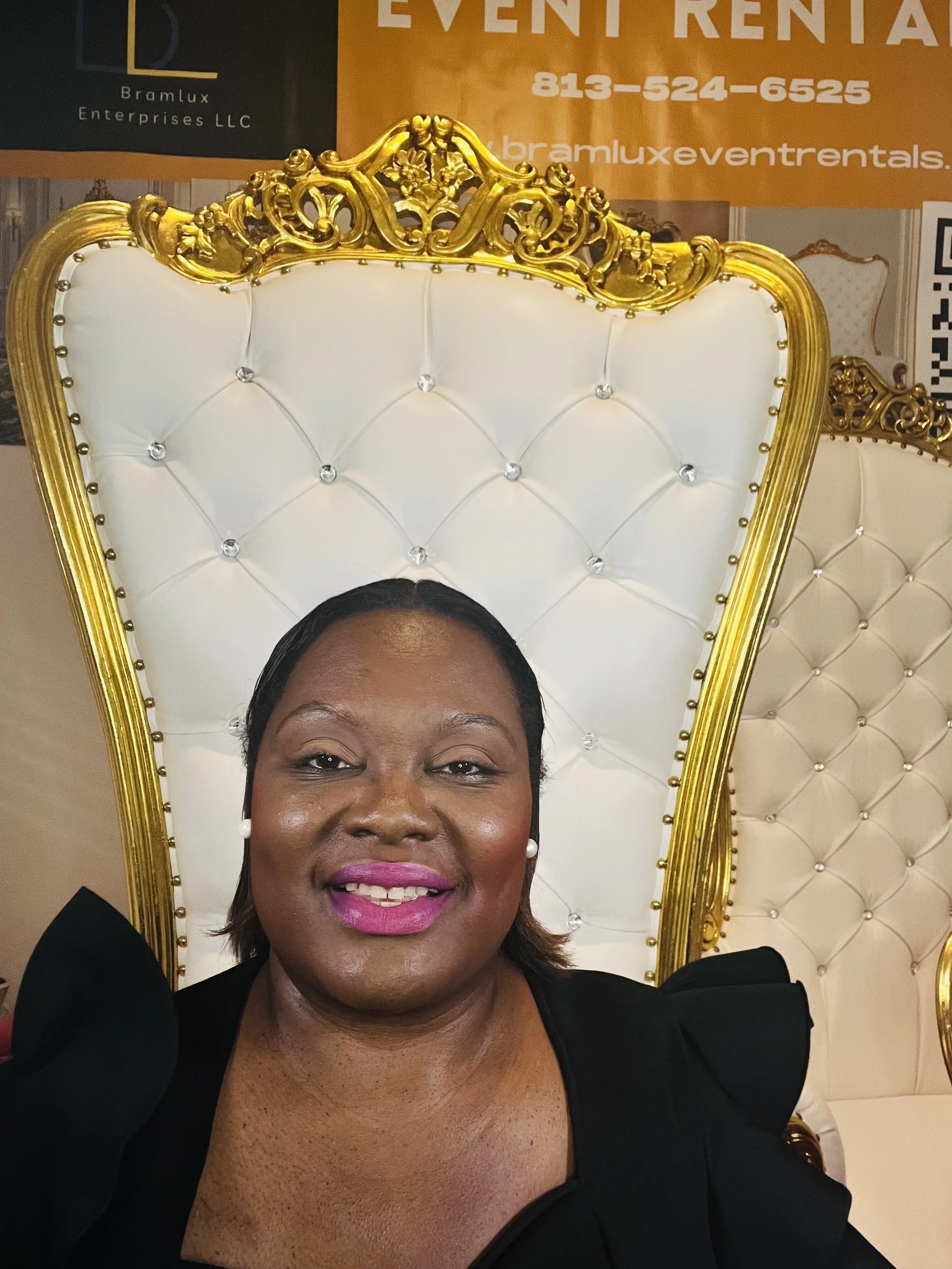 A woman with dark hair and pink lipstick sitting on a white and gold ornate throne-like chair, smiling at the camera.