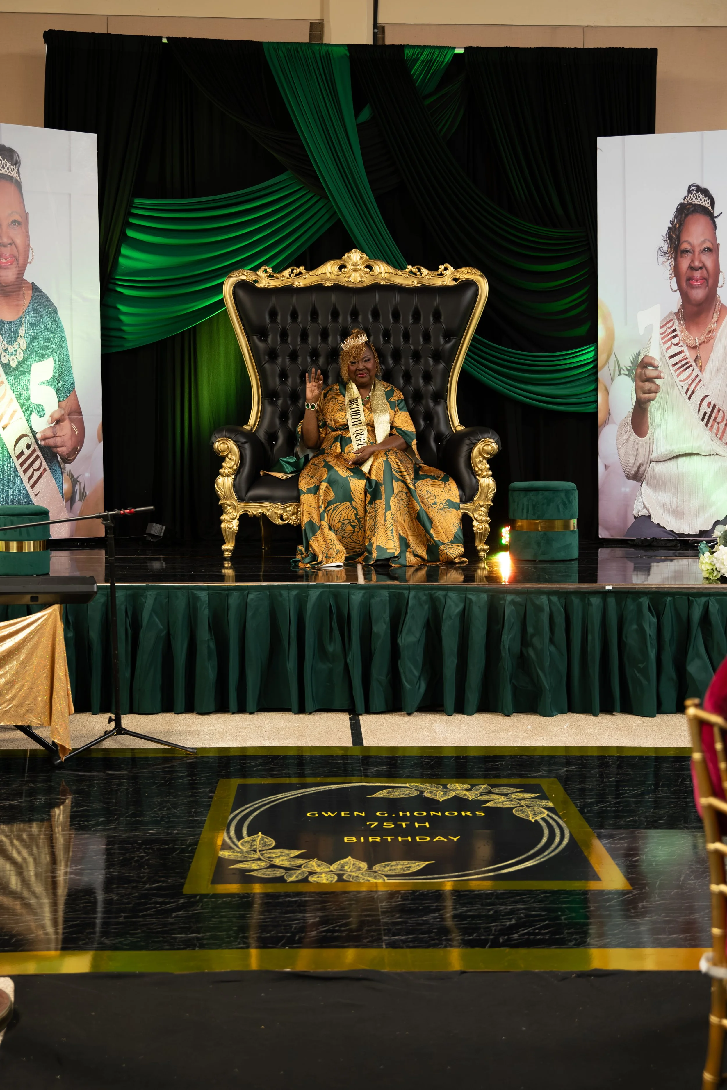 A woman wearing a crown and sash sits on a large, ornate black and gold throne on a decorated stage with green and black drapes, celebrating her 75th birthday. Banners with her photos and the words 'Gwen G. Honors 75th Birthday' are displayed around the stage.