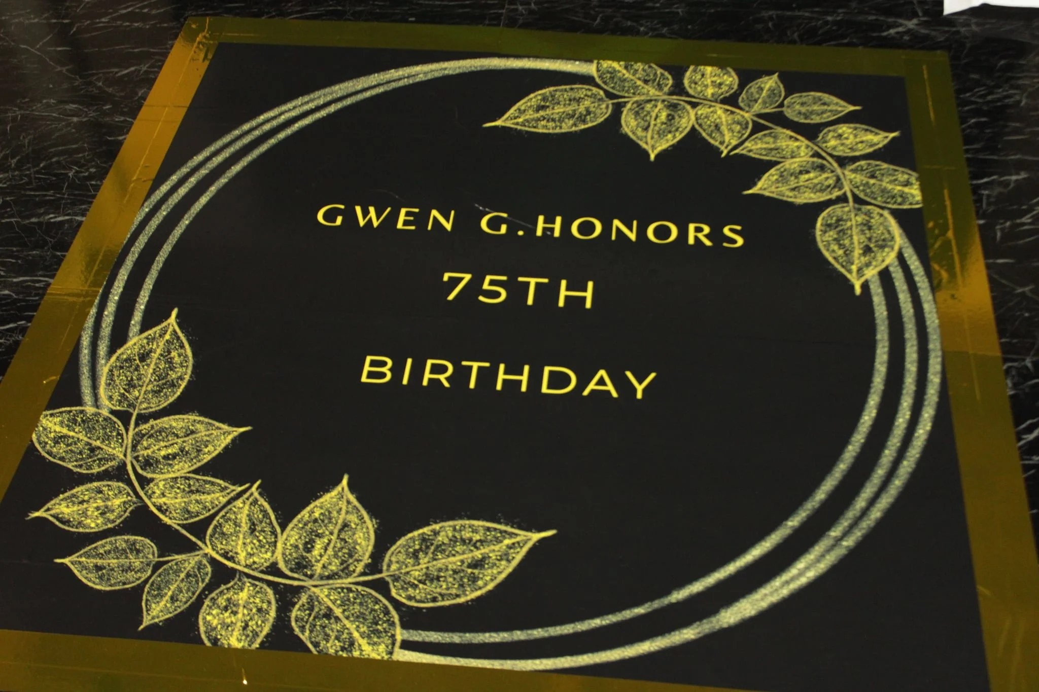 Black and gold birthday party sign with yellow lettering that reads 'Gwen G. Honors 75th Birthday' decorated with gold floral designs and circular borders.