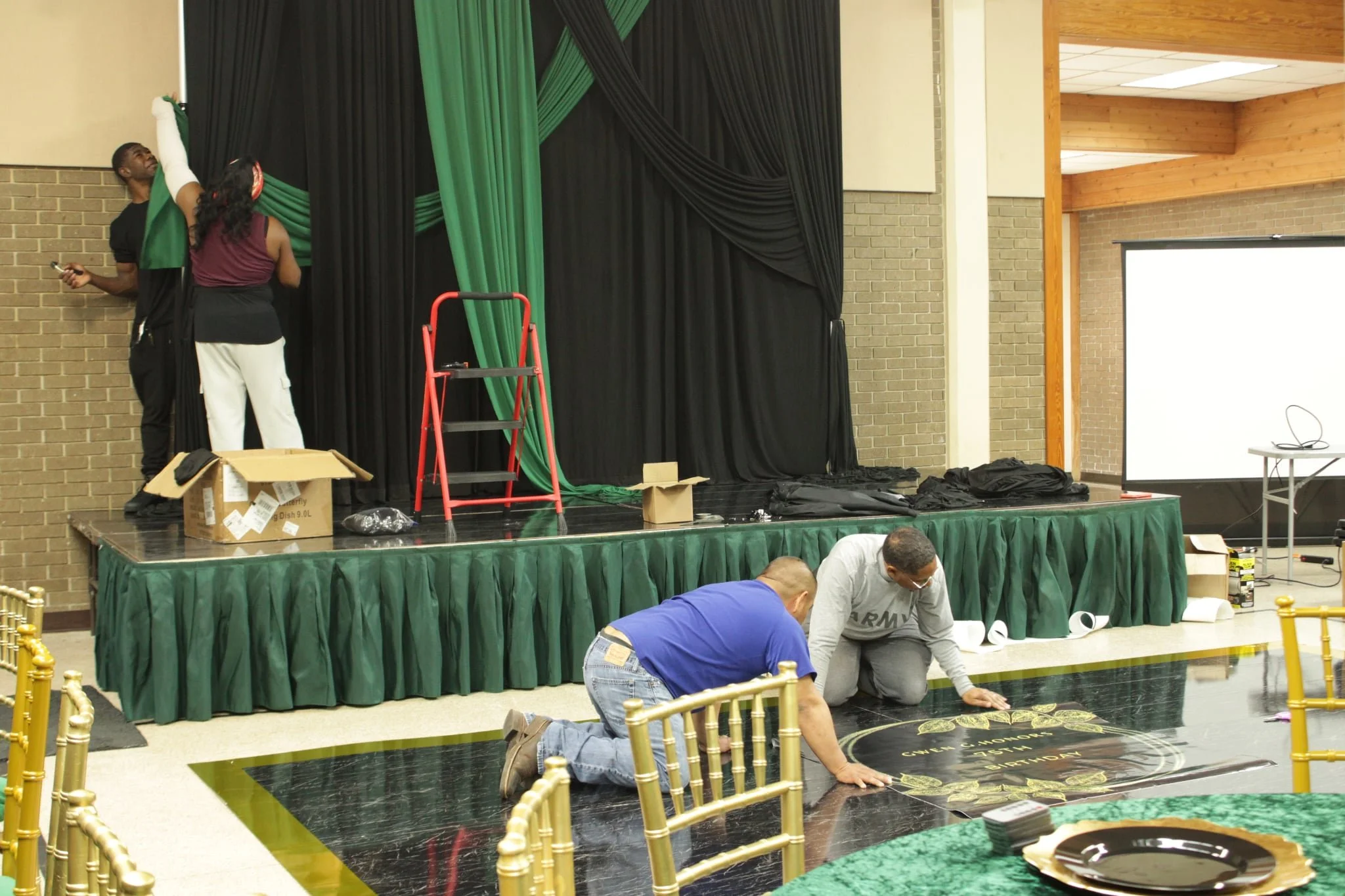 Two men kneeling on the floor installing a circular black and gold logo or sign on the floor, while three people stand on a stage behind them with black and green curtains and a ladder.