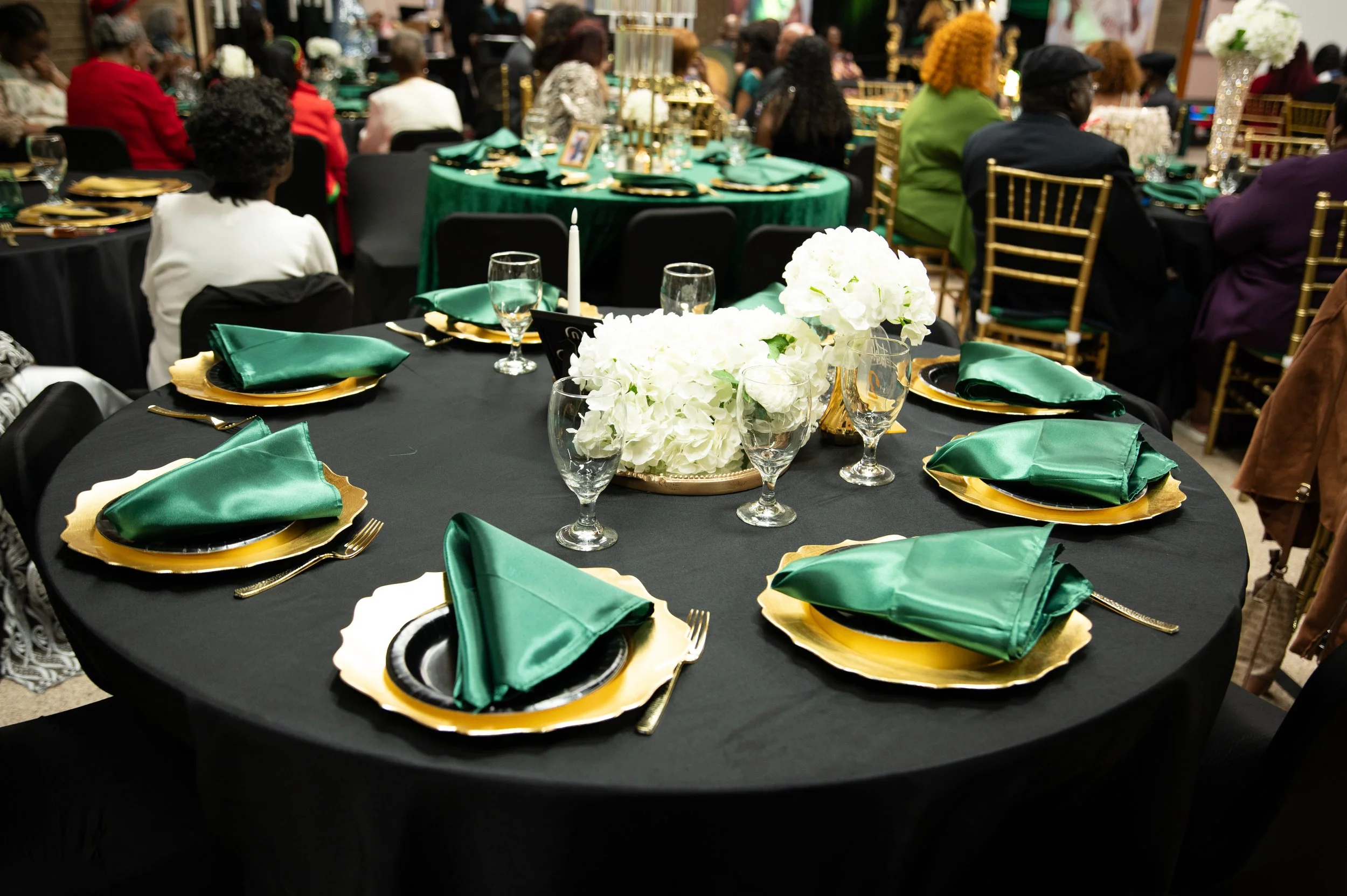 A banquet table with black tablecloth, green napkins, gold-rimmed plates, water glasses, and a centerpiece of white flowers, surrounded by guests at a formal event.