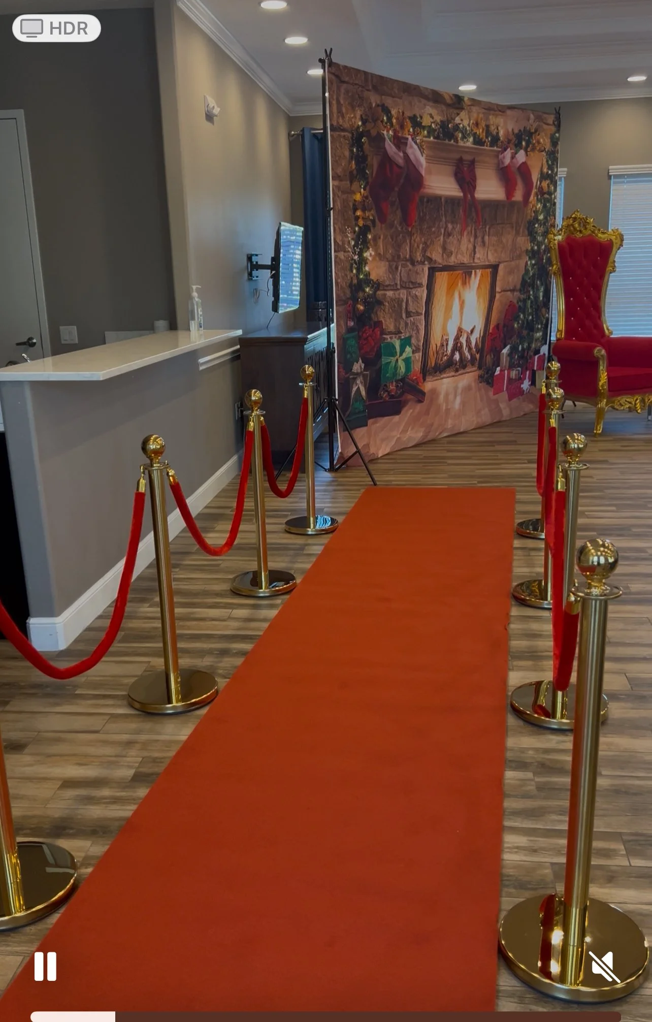 Empty red carpet on the floor leading to a fireplace backdrop with Christmas decorations and presents, surrounded by gold stanchions and red velvet ropes.