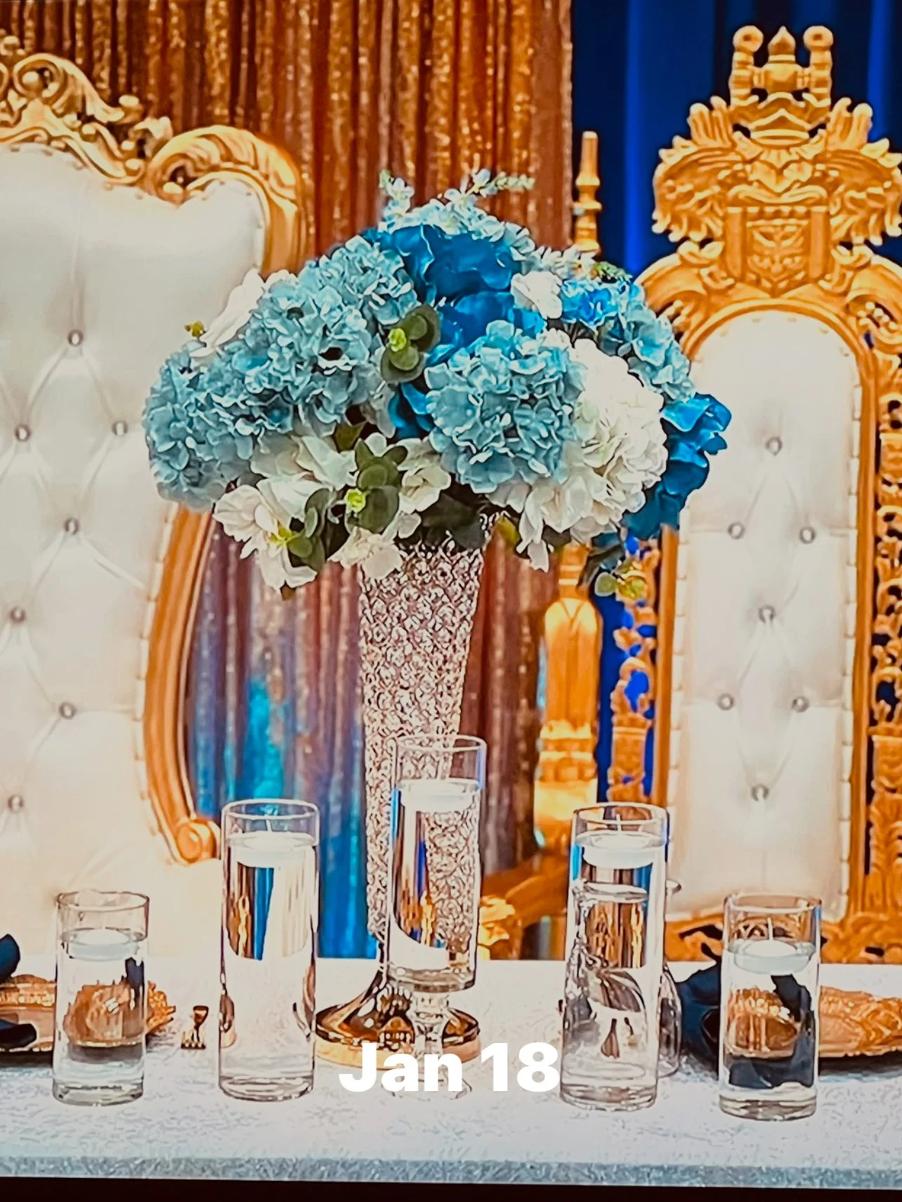 A tall, ornate silver vase with blue and white flowers, flanked by four glasses of water on a decorated table, with a luxurious, gold-accented white and cream chair and a blue curtain in the background.