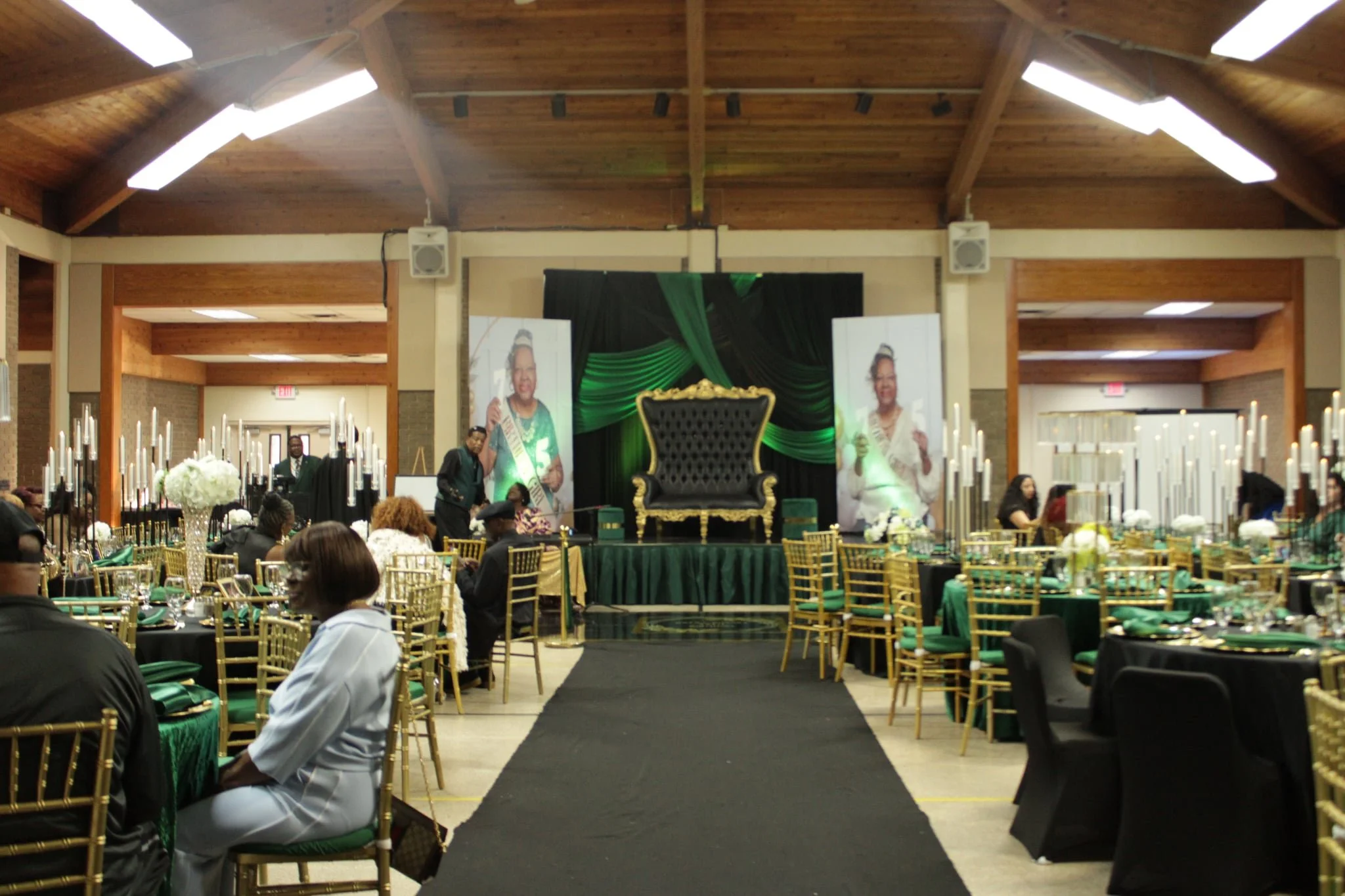 Event hall decorated with black and green drapes, gold chairs, and floral centerpieces, featuring a stage with a large black and gold throne-style chair and two large portraits of women in traditional attire.