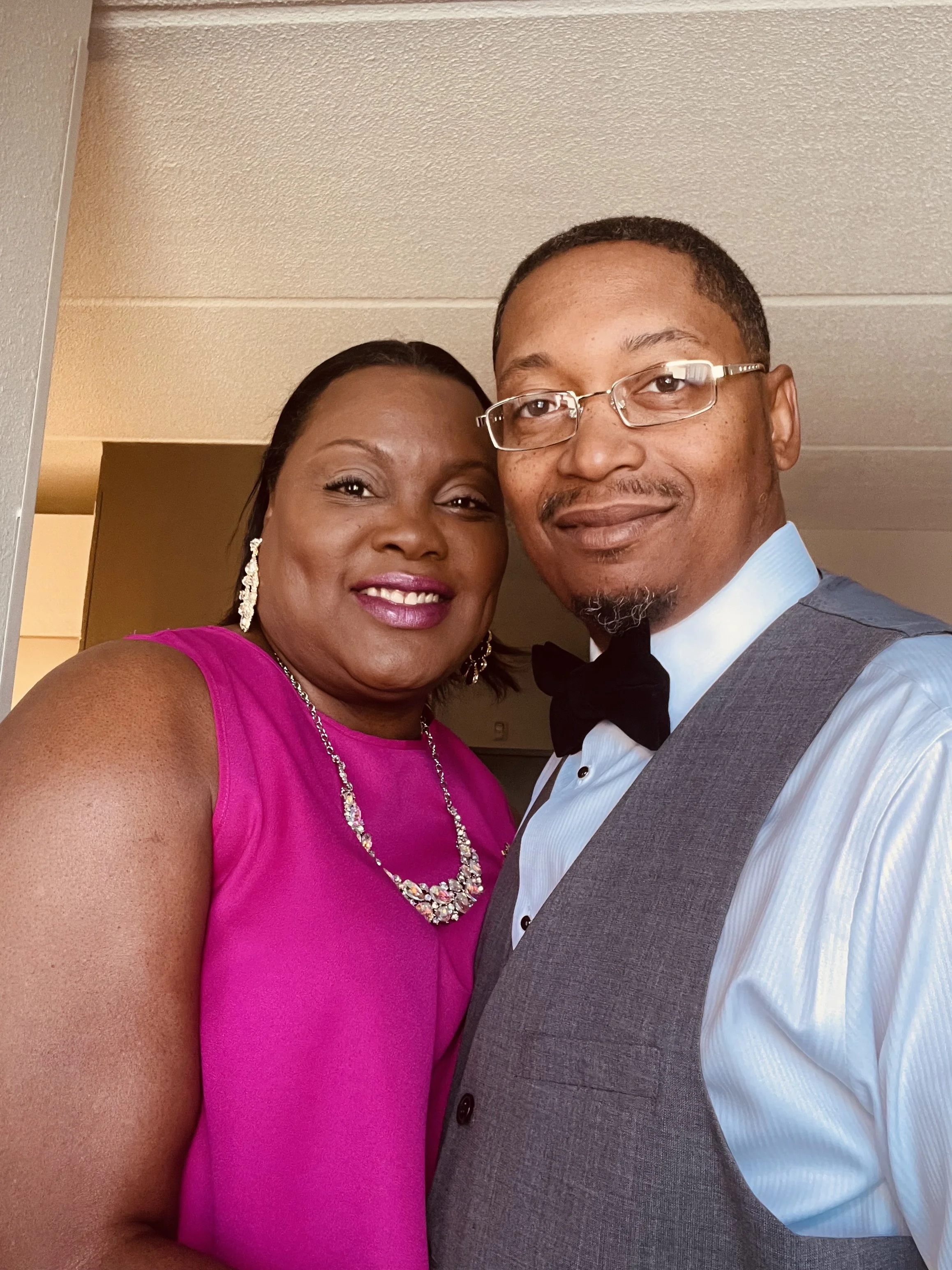 A smiling woman in a pink dress and a man in a tuxedo with glasses take a selfie together indoors.