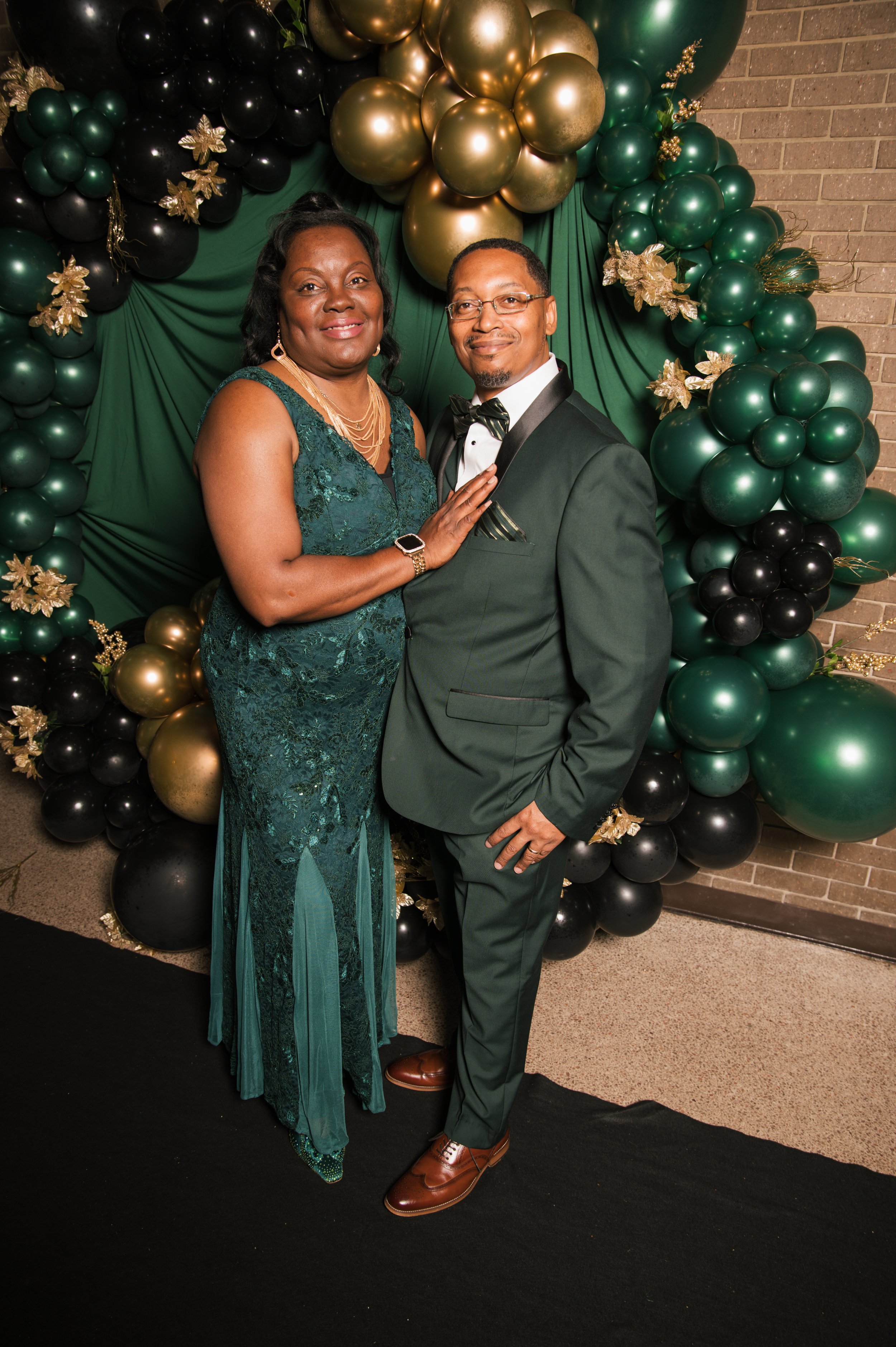 A smiling woman in a teal lace gown and a man in a dark suit with a bow tie, standing side by side in front of a green, black, and gold balloon backdrop with gold floral accents.