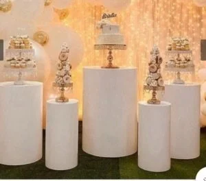 Elegant dessert table with 3 tall white pedestals displaying layered desserts and a birthday cake in the center, against a gold and peach backdrop.