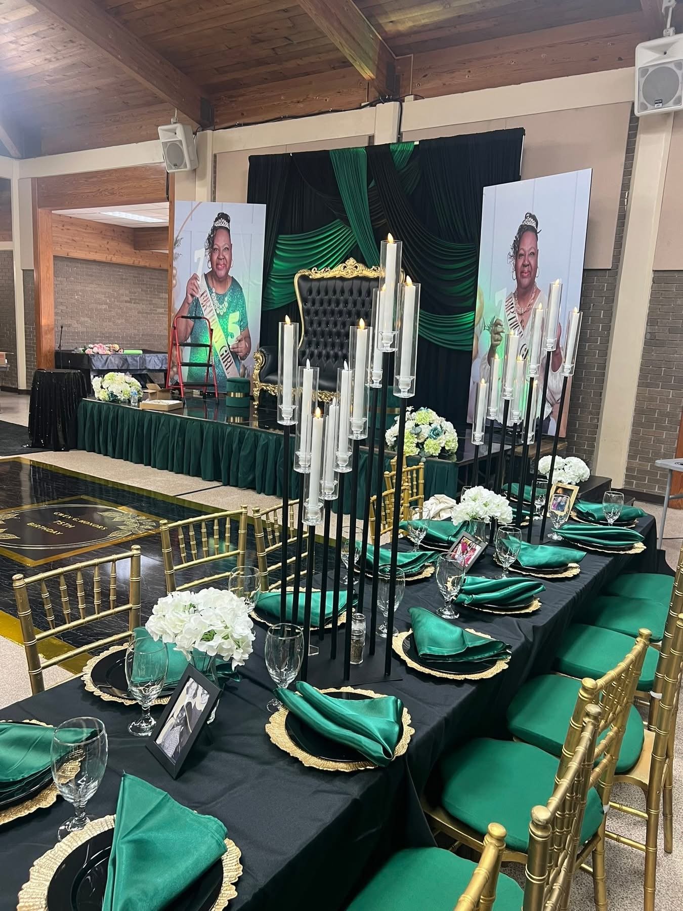 A decorated event space with a long table set with green napkins, glasses, and floral centerpieces, and a stage featuring large portraits of a woman wearing a crown and green dress, with black and green drapery backdrop and tall white candles.