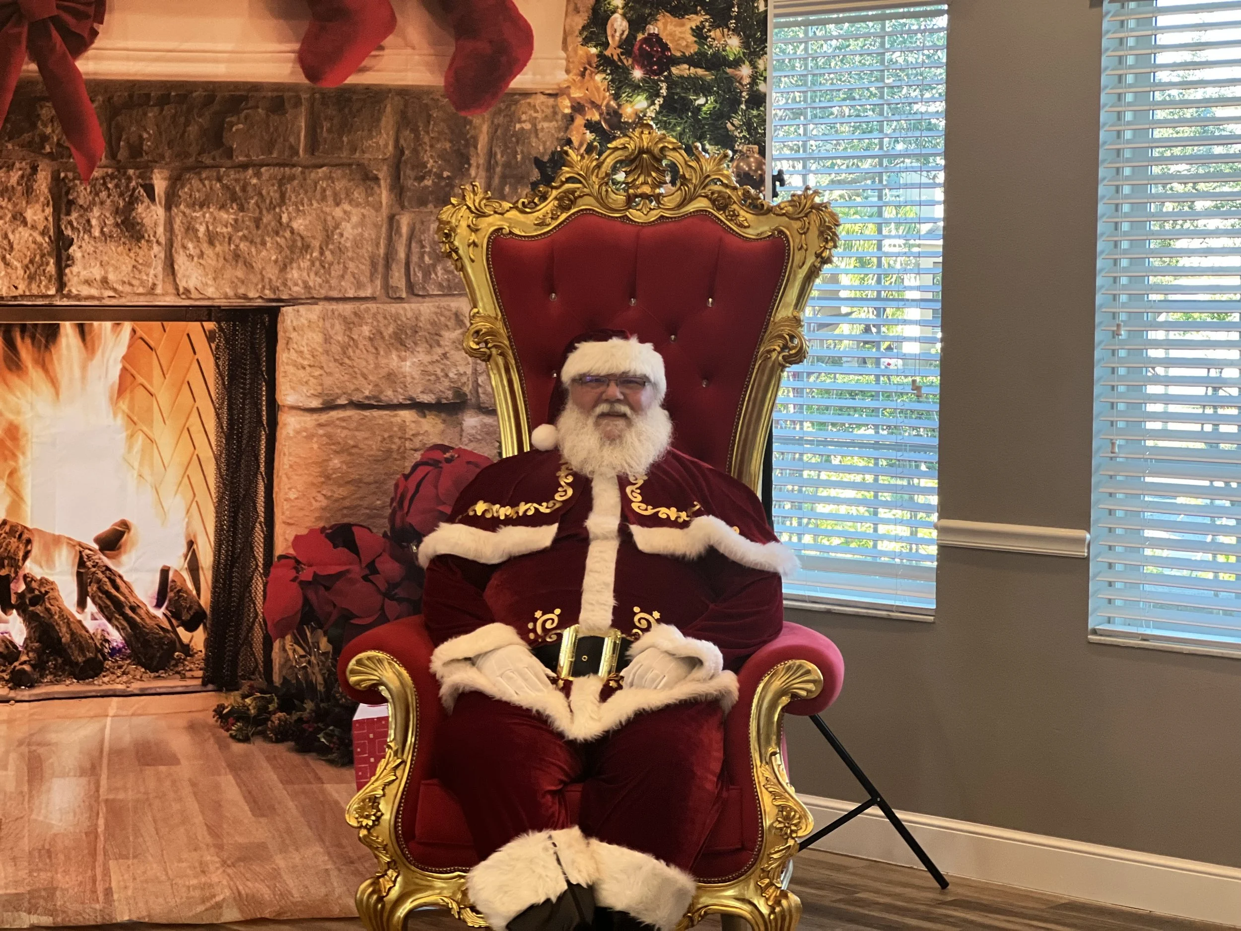 Santa Claus with a white beard and glasses sitting on a red and gold ornate throne in front of a fireplace with stockings and a Christmas tree, beside a window with blinds.