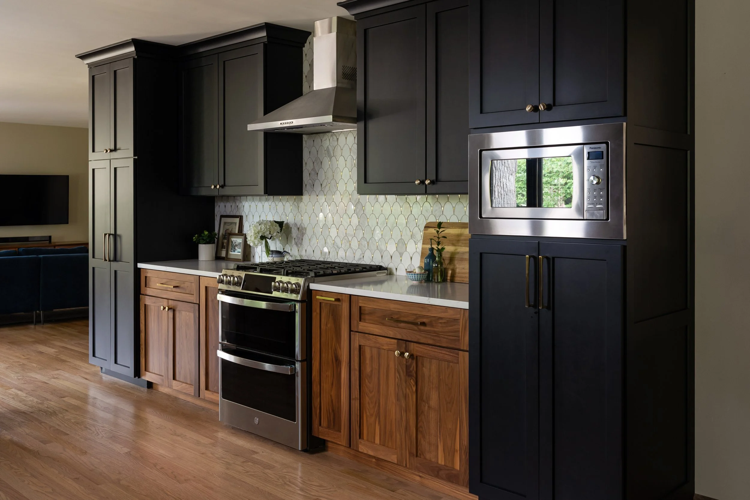 A modern kitchen with black oak and natural walnut cabinetry, stainless steel appliances, a gas stove, decorative backsplash tiles, and wooden flooring.