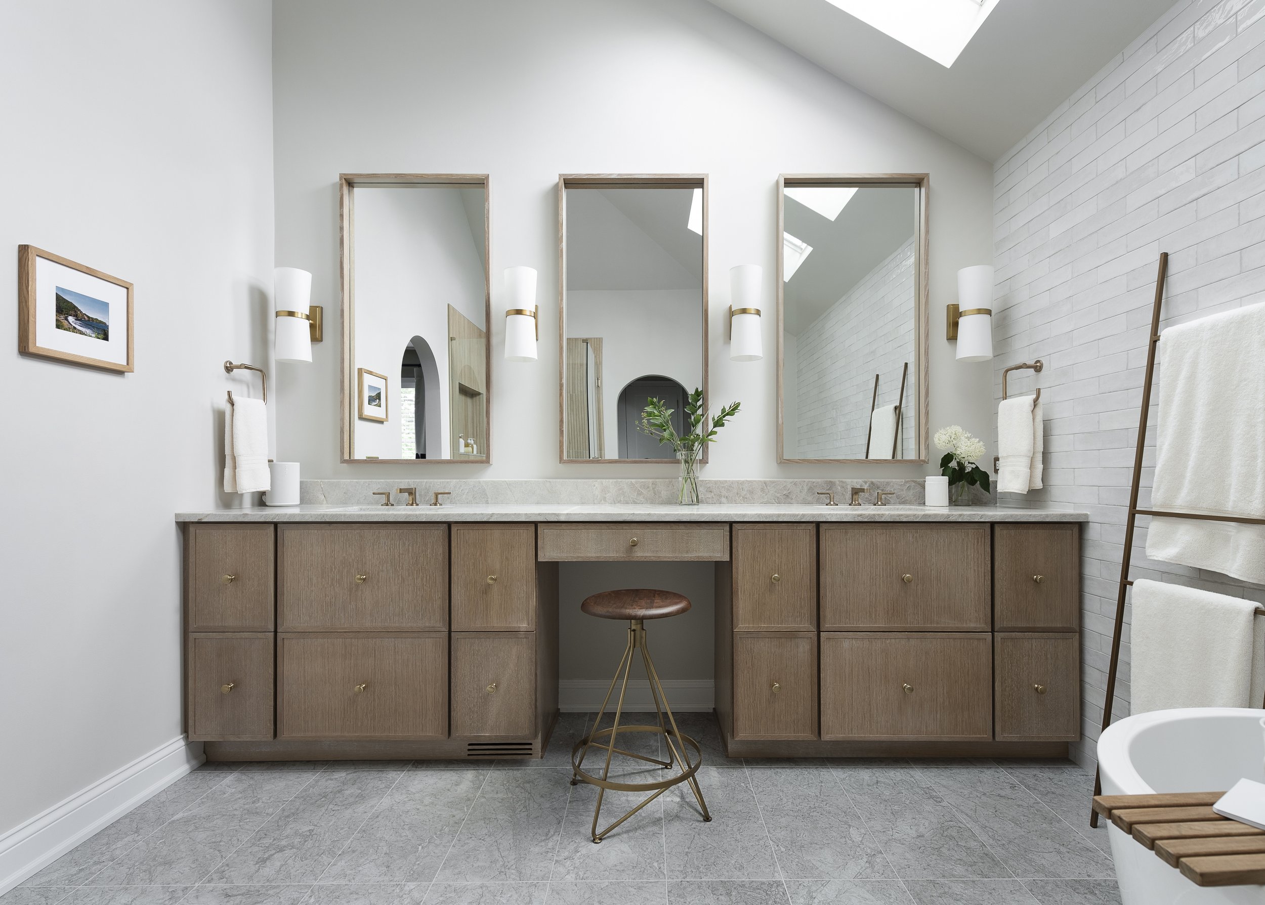 Modern bathroom with double vanity, three mirrors, white tiled wall, ladder towel rack with white towels, light gray floor, and a white tub with a wooden tray holding a cup.