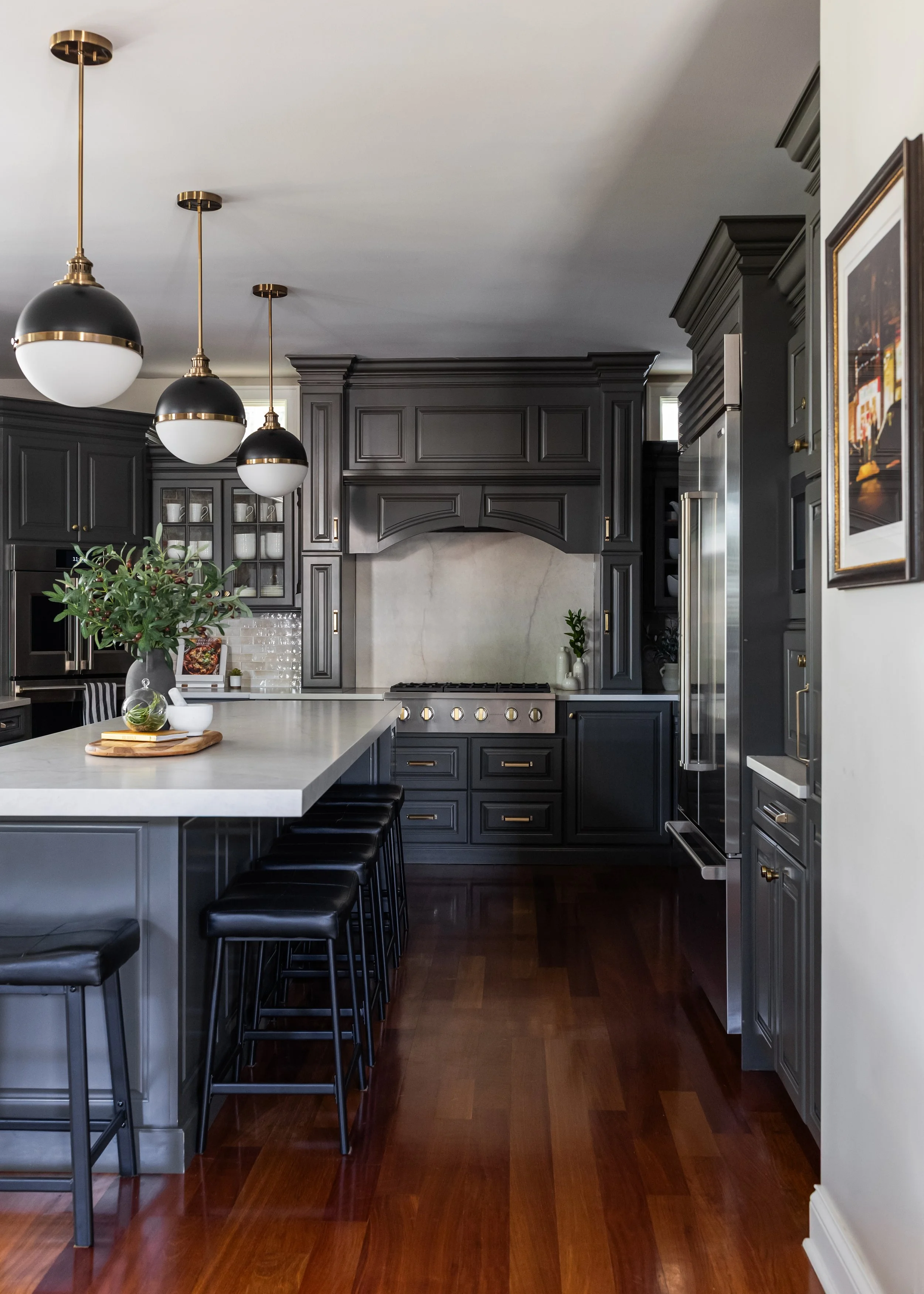 A transitional kitchen with dark gray taupe cabinets, an ivory island with black stools, a stainless steel refrigerator, a range hood, pendant lights, and hardwood flooring.