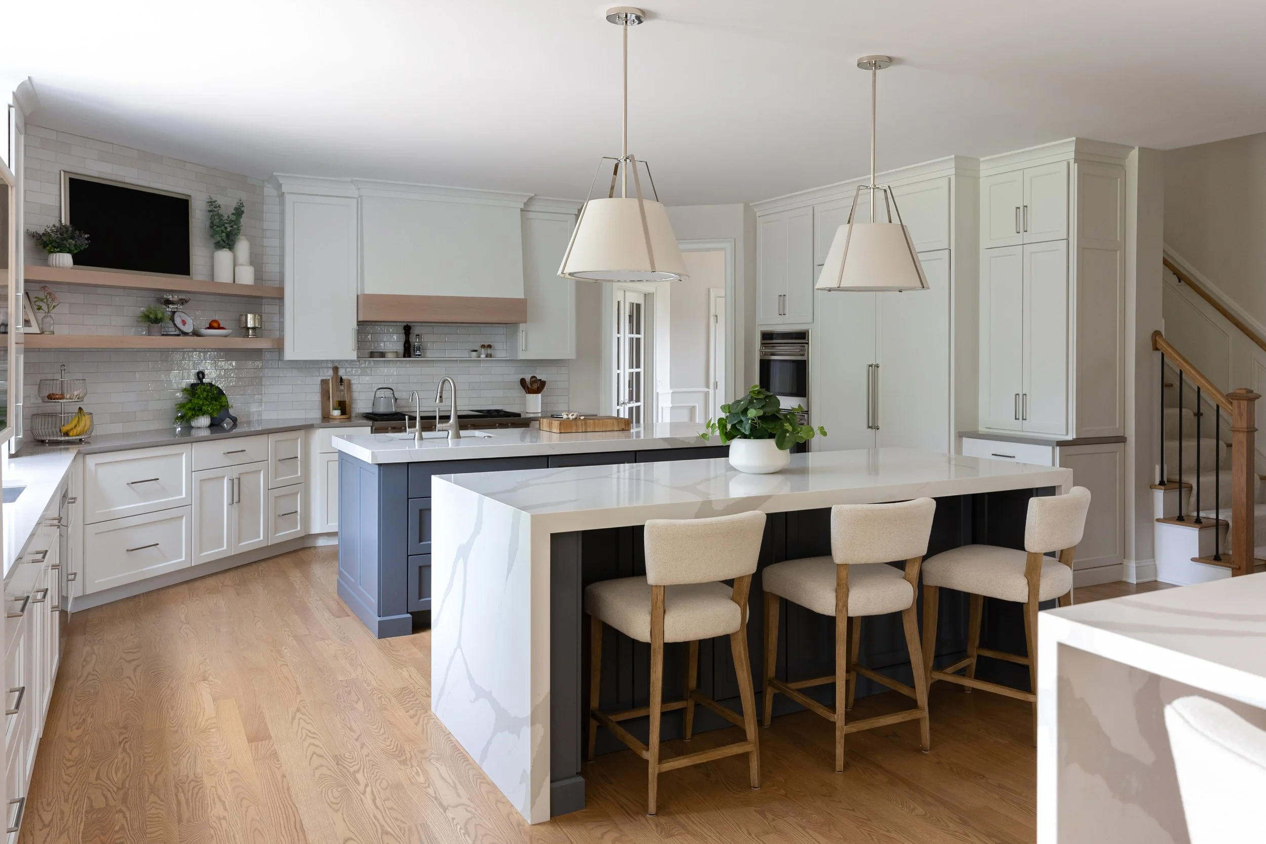 Modern white kitchen with island and beige bar stools, pendant lights, white cabinets, and wooden flooring.