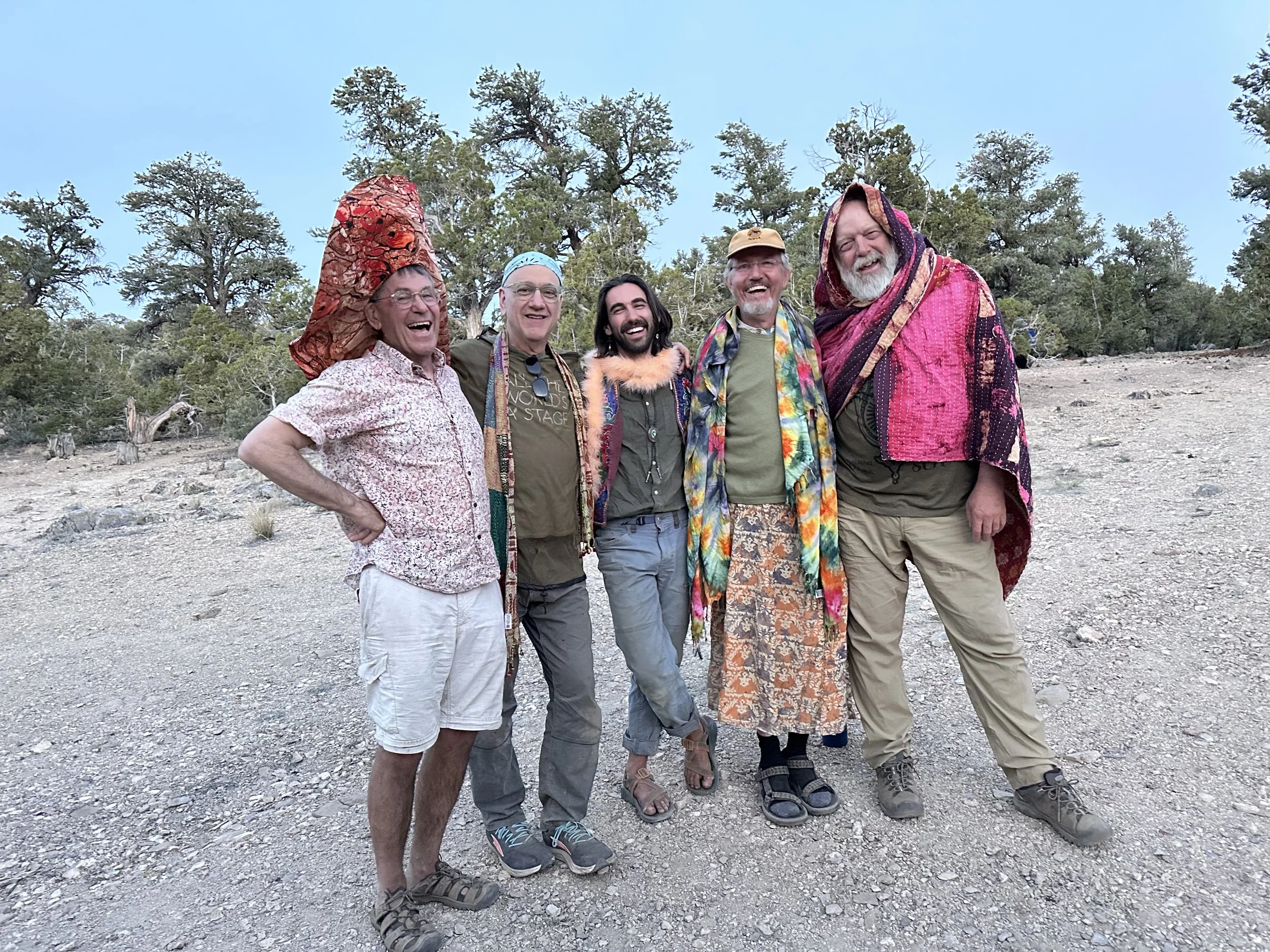 Five queer men, the guides for an outdoor program, in the Inyo mountains with trees in the background, dressed in casual, playful, and colorful clothing, smiling and laughing together.