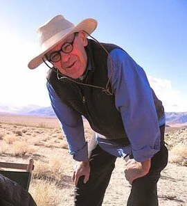 Older man in sunglasses and a wide-brimmed hat smiling outdoors in a desert landscape