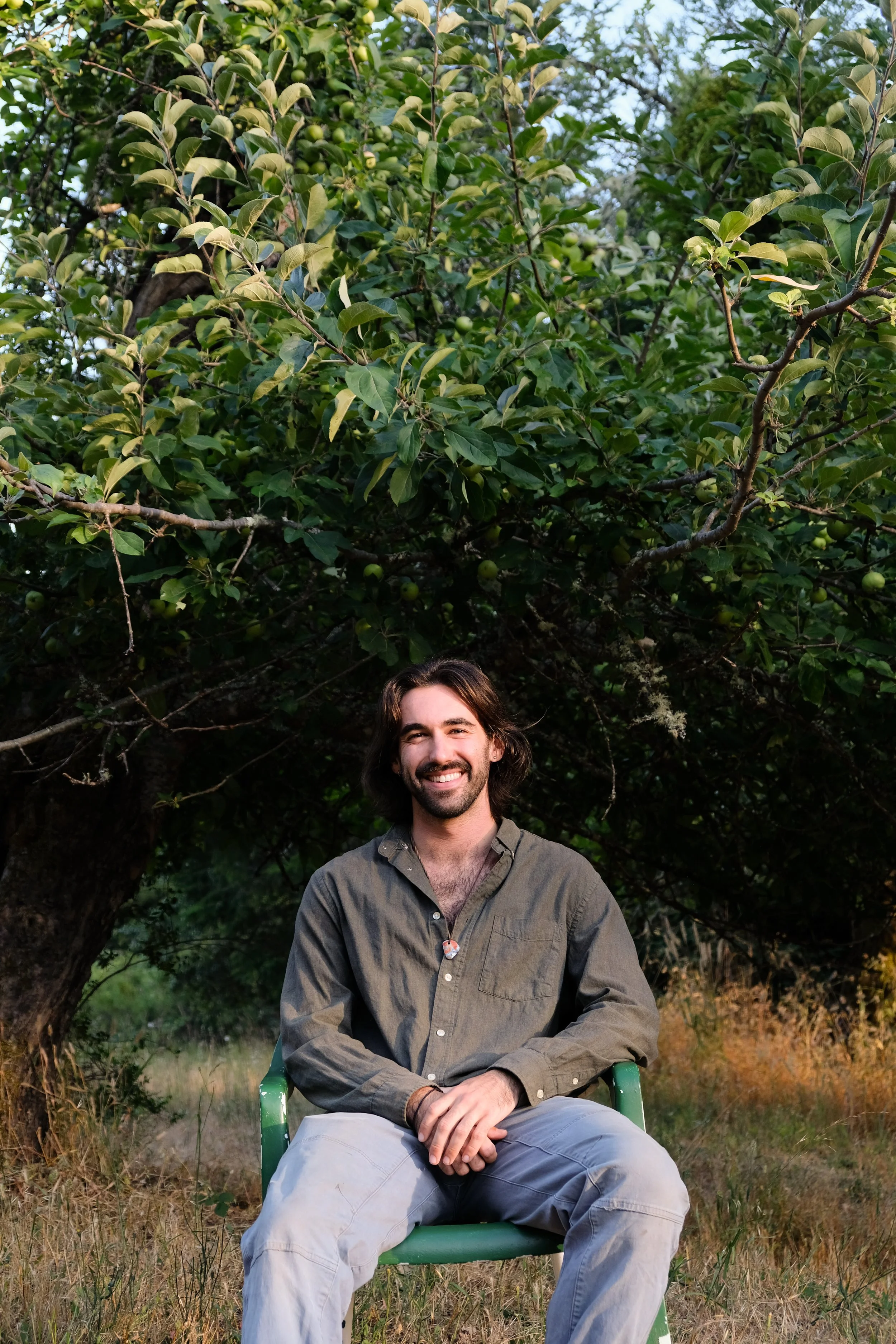 A man with long dark hair and a beard sitting on a green chair outdoors in front of a large leafy tree, smiling at the camera.